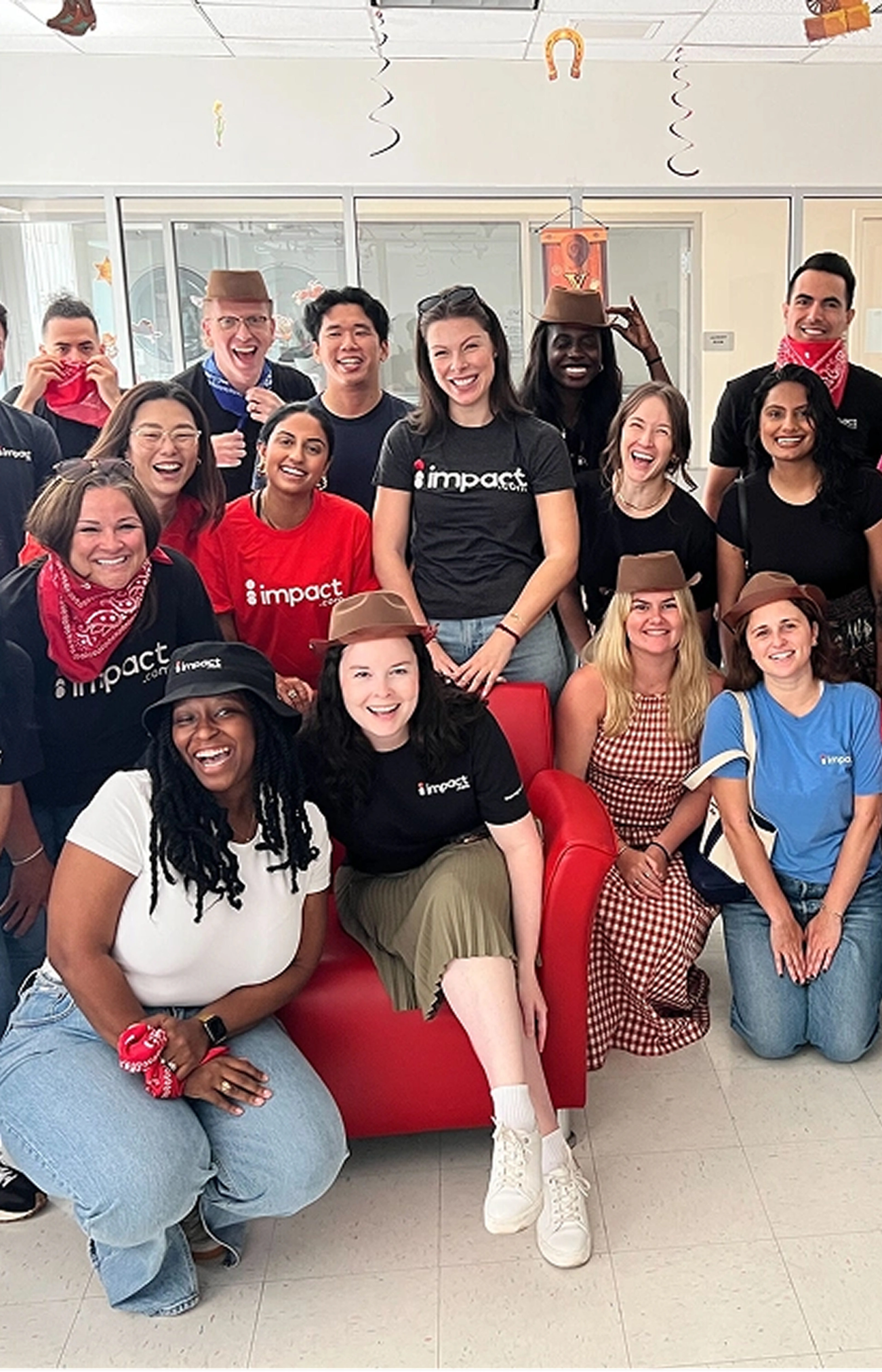 A group of smiling people wearing impact shirts, red bandanas, and brown paper hats pose together indoors around a red chair; some are sitting while others stand behind them.