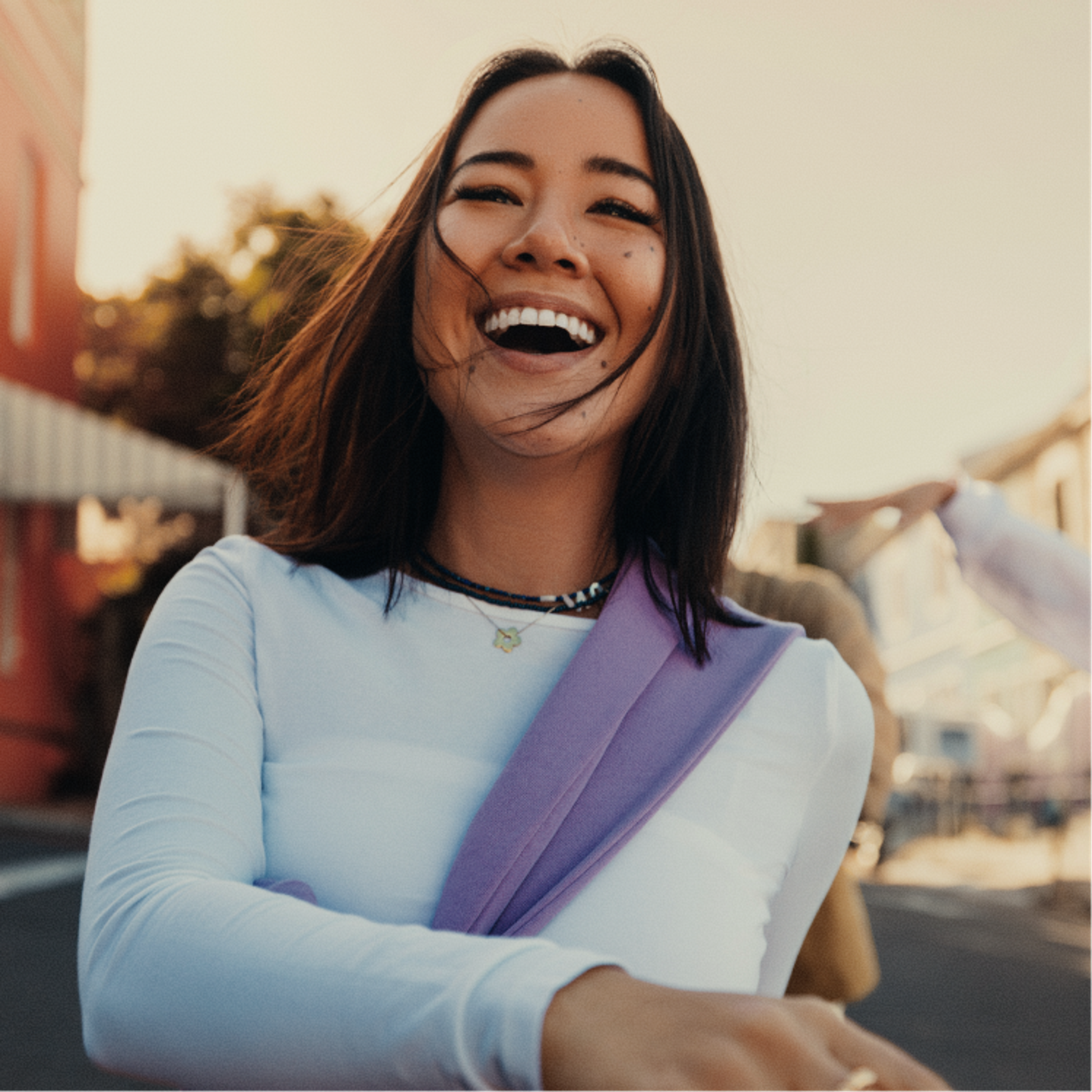 Young woman with straight dark hair, wearing a white shirt and lavender scarf, smiles brightly outdoors on a sunny day. A blurred street and buildings are in the background.