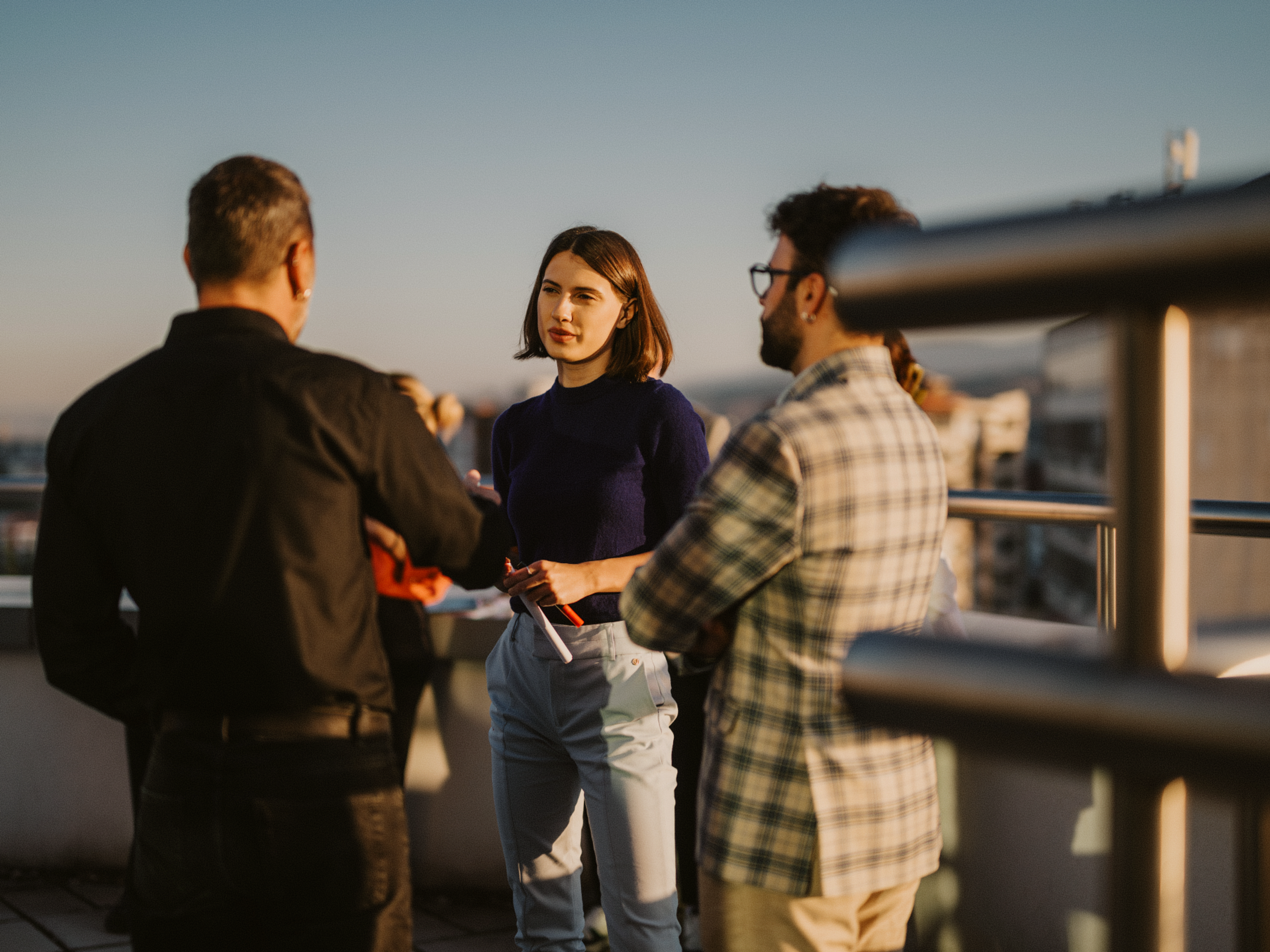 Three colleagues talking on a rooftop terrace at sunset.