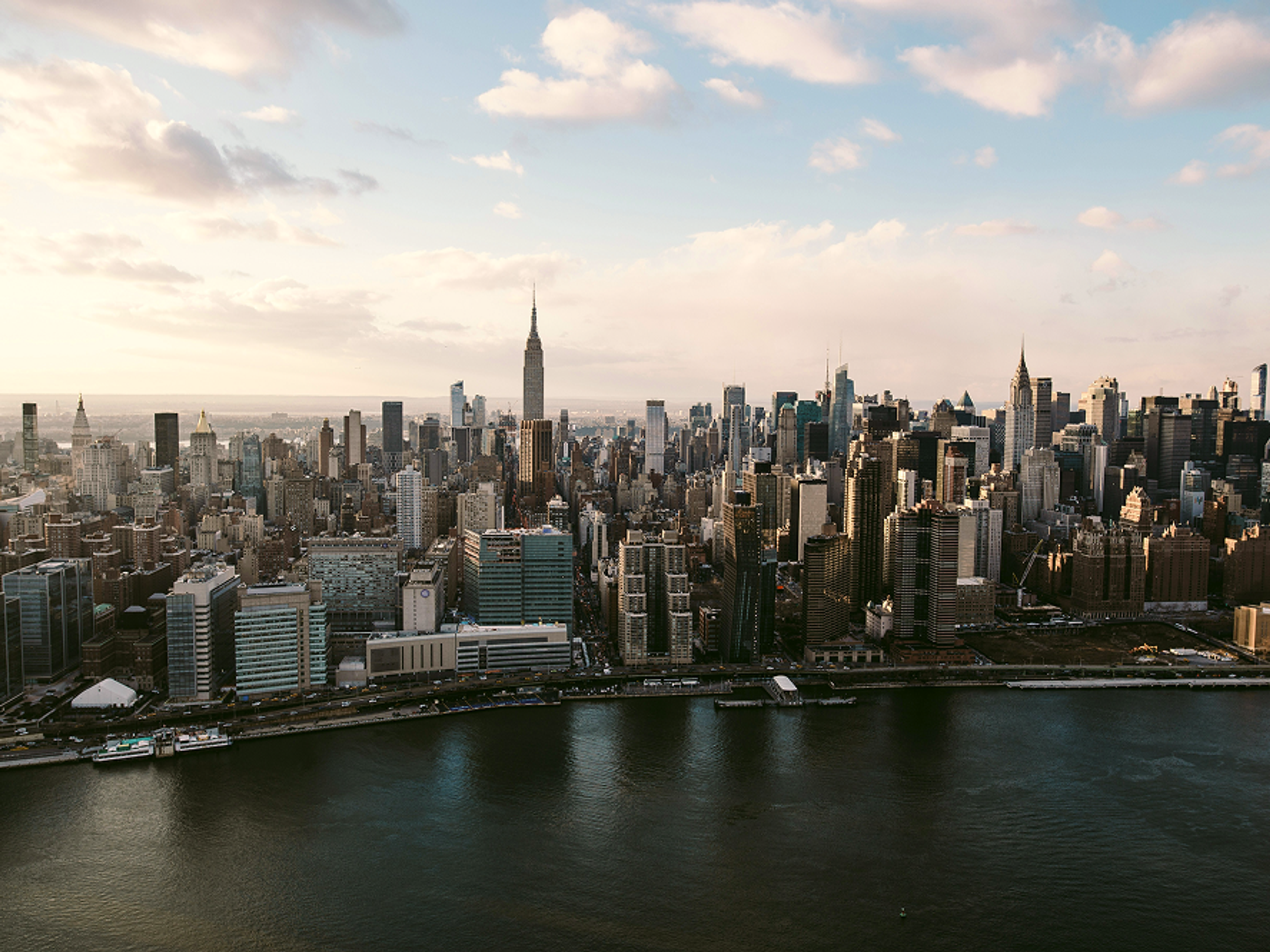 A view of Manhattans skyline at sunset, with skyscrapers and the Empire State Building visible, seen from across the East River. The sky is partly cloudy.