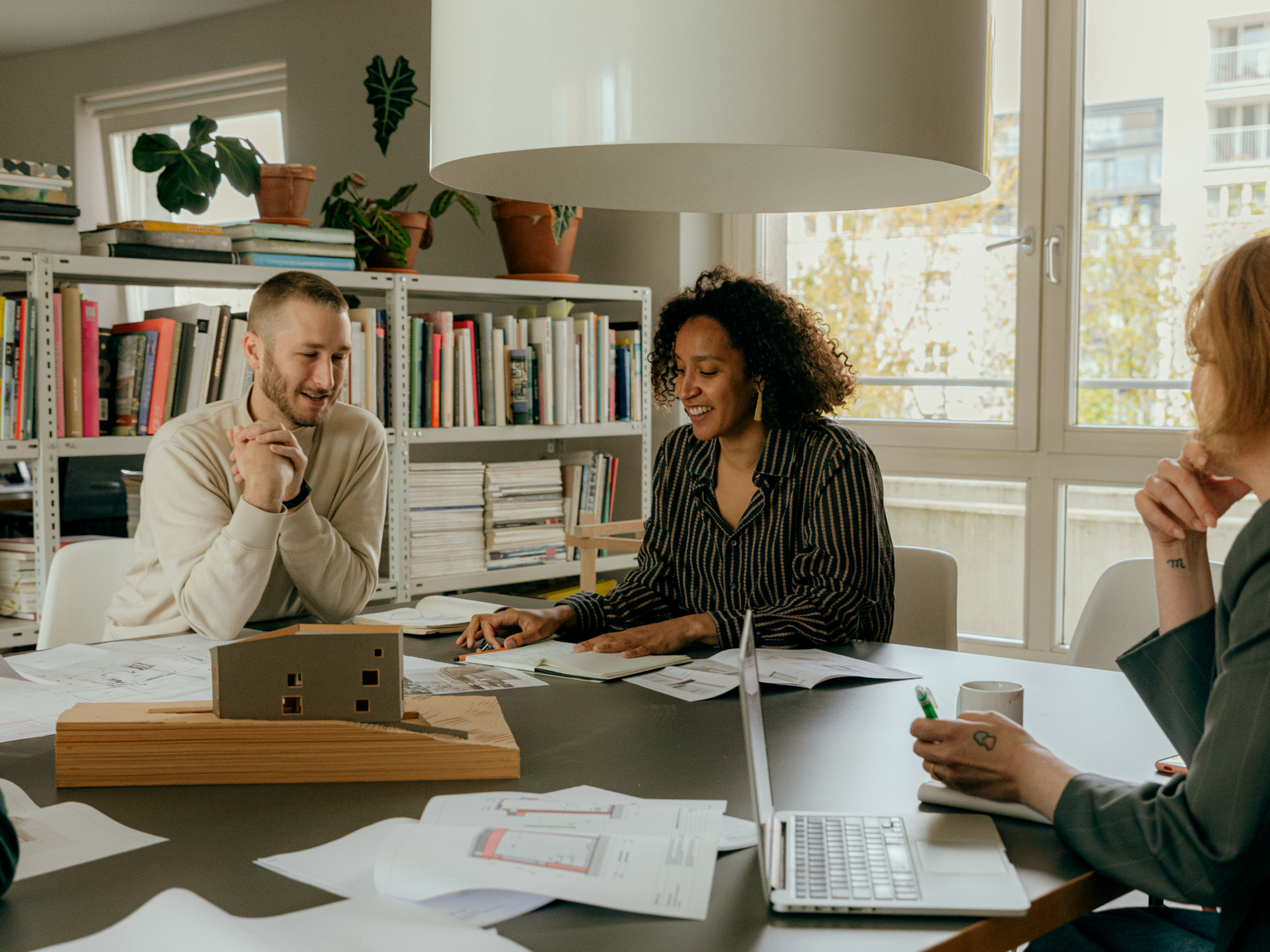 Three people sit around a table with papers, a laptop, and a building model, discussing ideas in a modern, bright office with bookshelves and plants in the background.