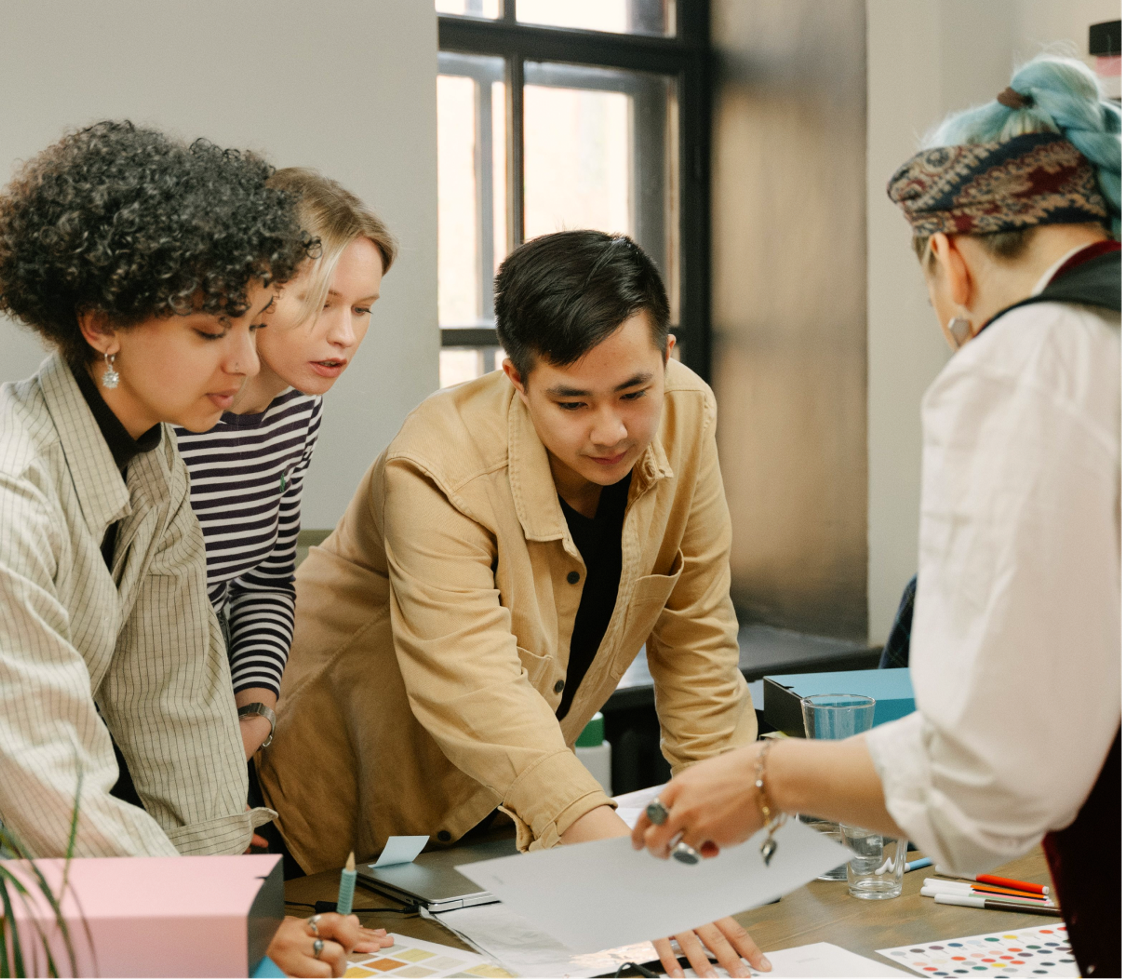 Four people collaborate at a desk, looking at papers and color swatches. One person gestures while the others lean in, engaged in discussion in a bright, modern office.