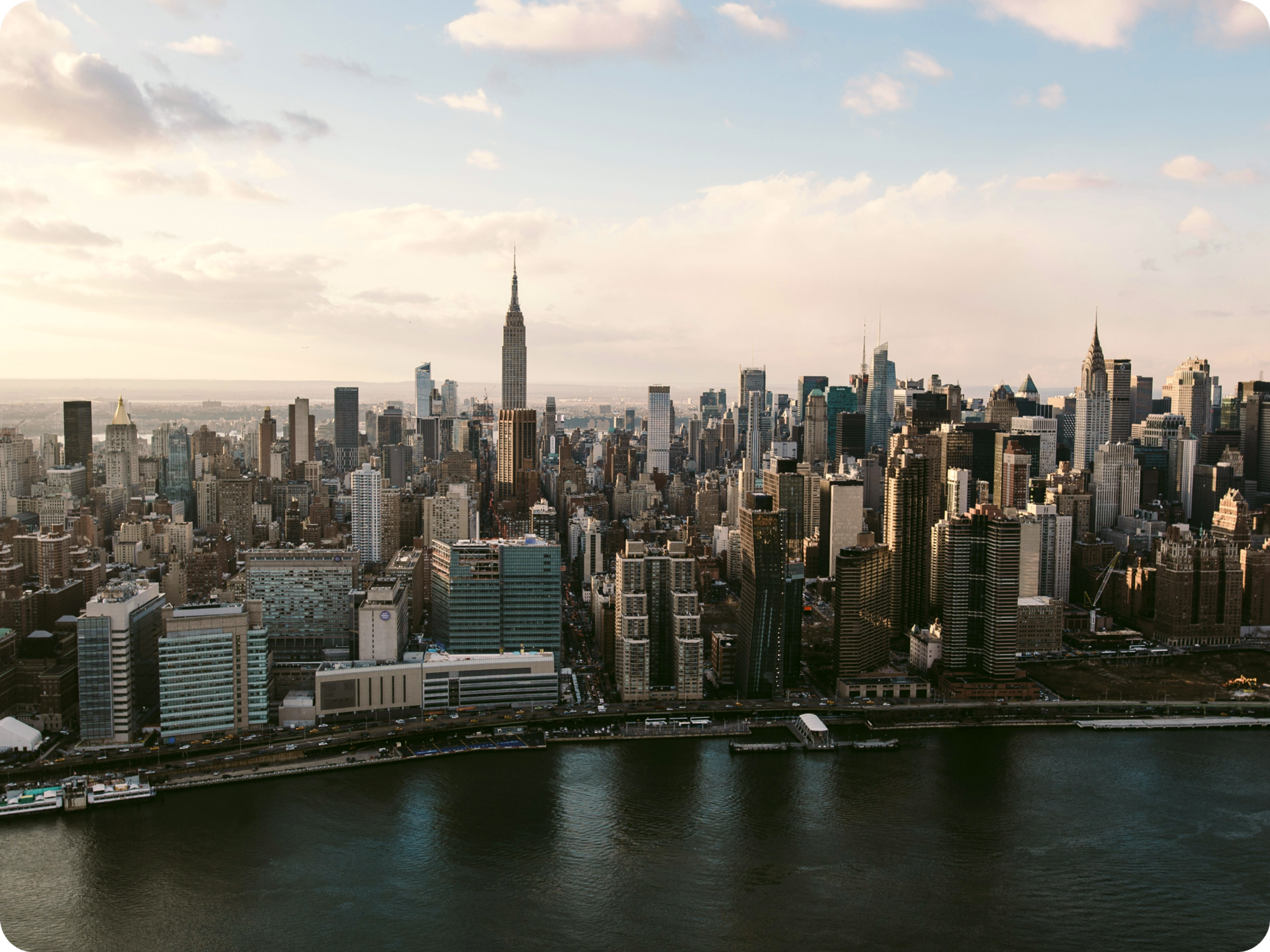 Aerial view of New York City skyline with the Empire State Building centered, skyscrapers surrounding it, and the East River in the foreground under a partly cloudy sky.