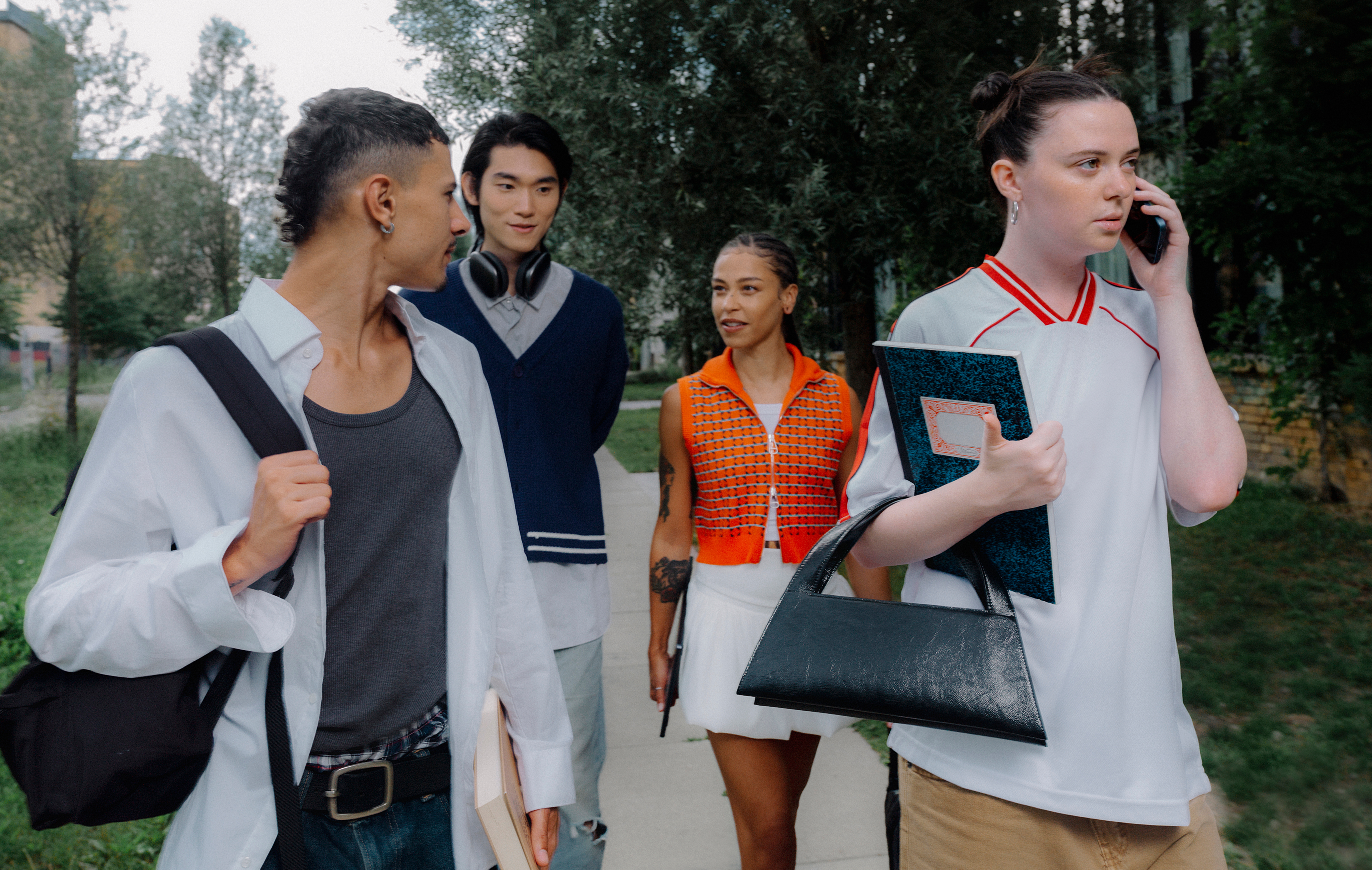 Four young adults walk outdoors. One holds a phone to their ear, while others chat and carry bags or notebooks. Trees and buildings are in the background.