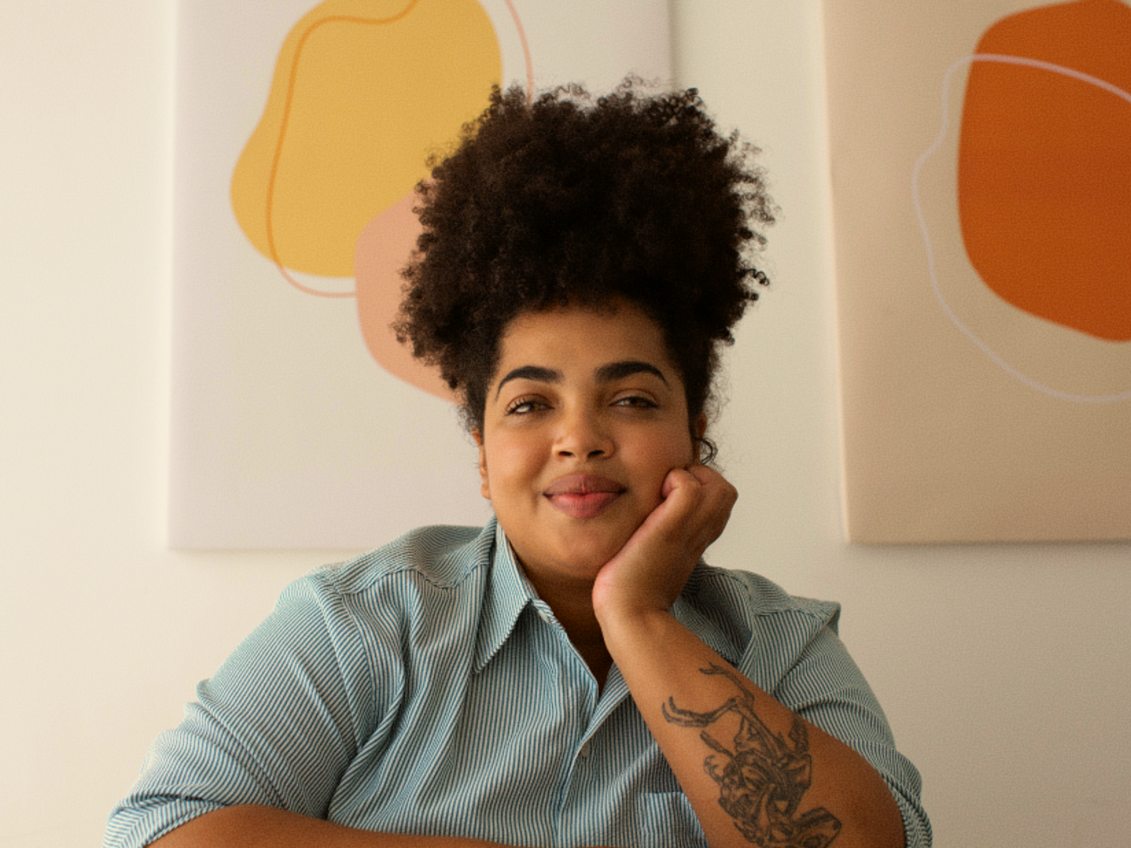 A person with curly hair and a tattoo on their forearm smiles gently, resting their chin on their hand. Abstract art hangs on the wall in the background.