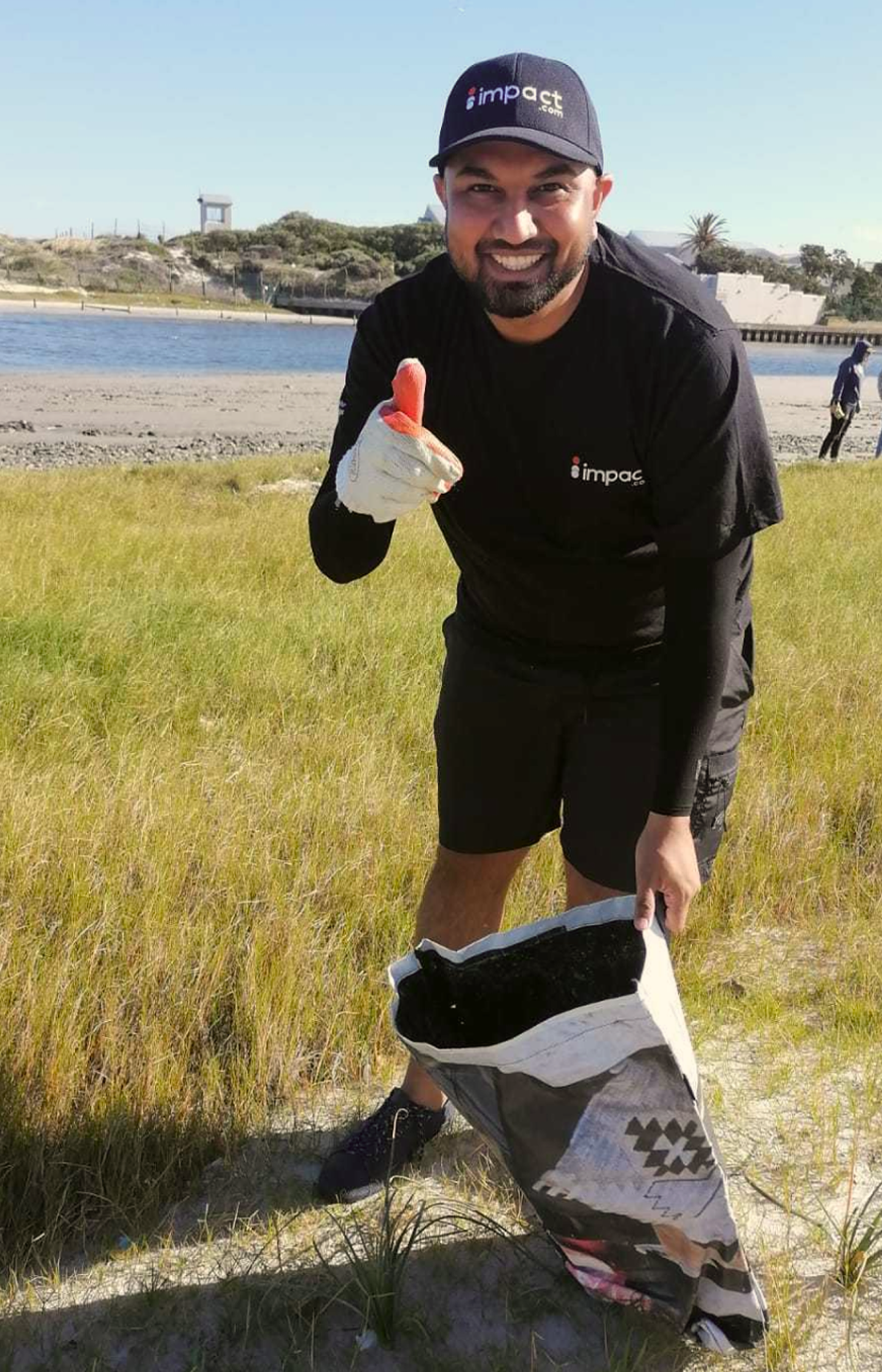 A man wearing gloves and an impact shirt picks up litter on a grassy beach, smiling and holding a piece of trash, with a bag and water in the background.