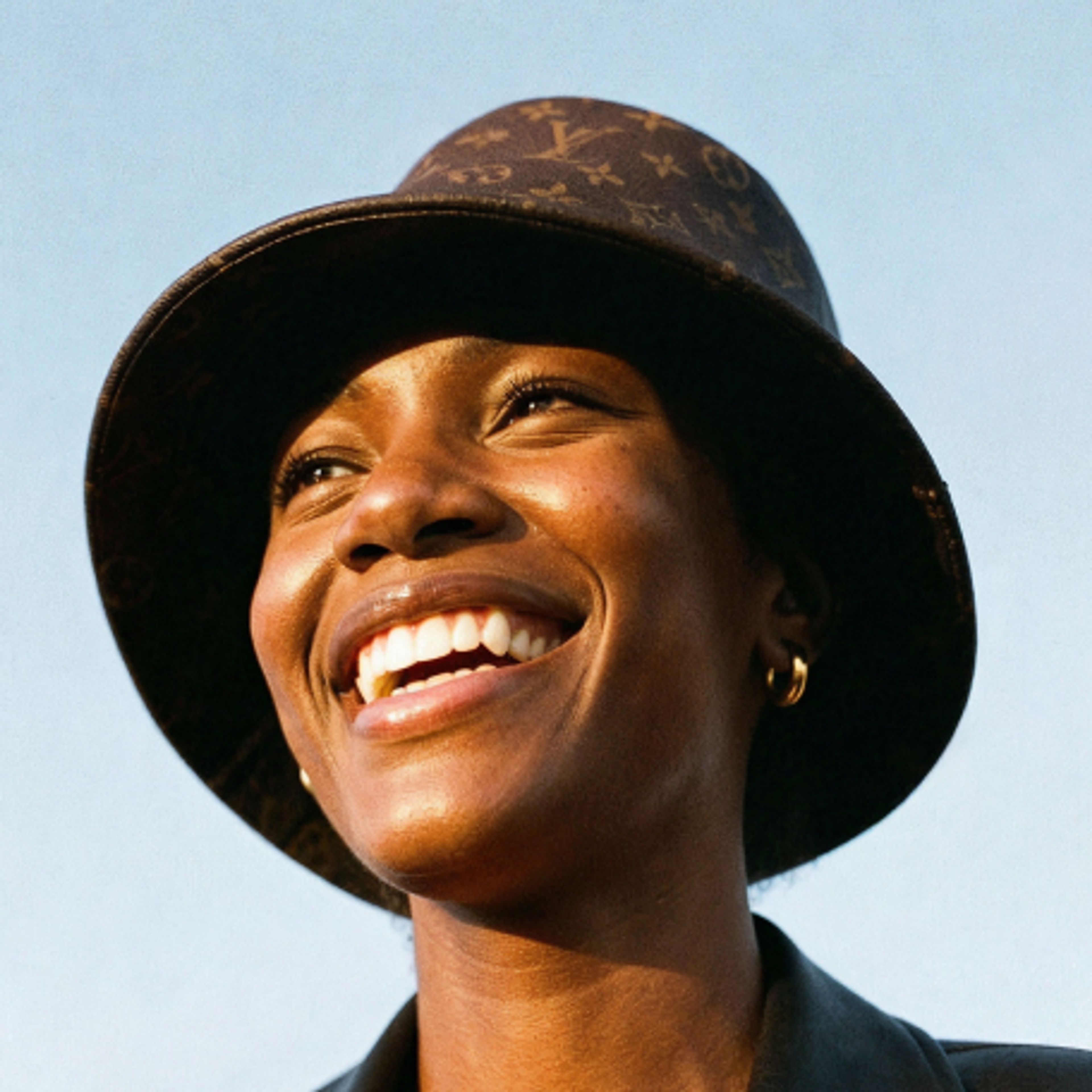 A person smiling brightly, wearing a dark bucket hat with a subtle pattern and gold hoop earrings, photographed against a clear blue sky.