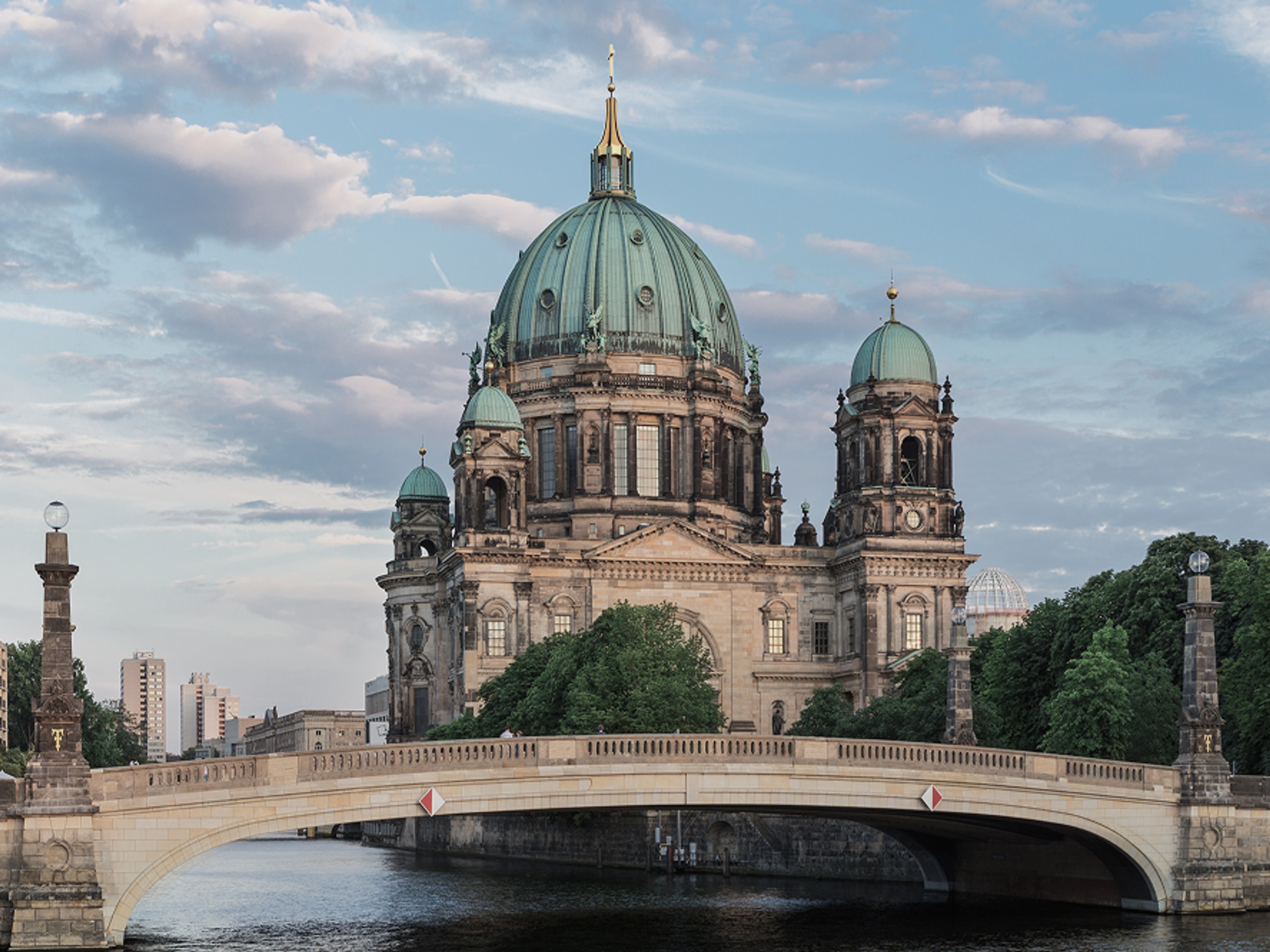 A large domed cathedral, the Berlin Cathedral, stands behind a stone bridge over a river, with lush trees and buildings in the background under a partly cloudy sky.