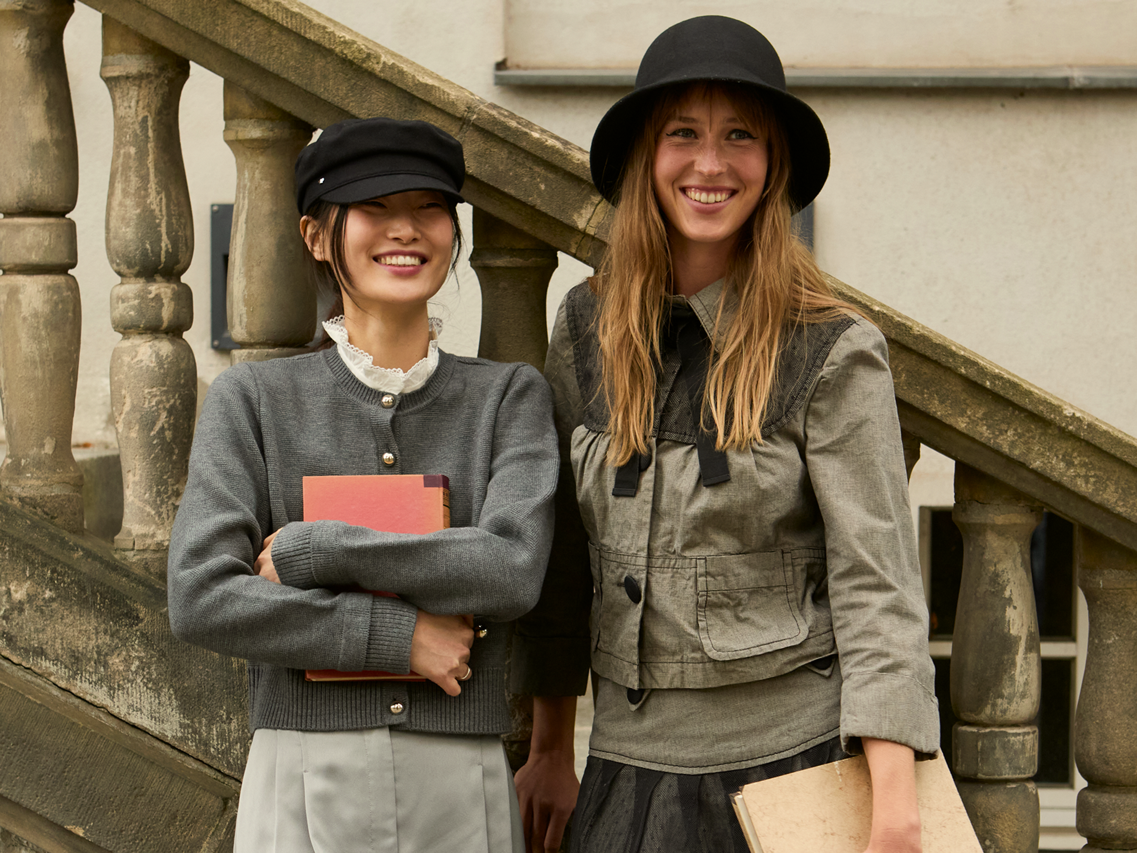 Two women stand outside by stone stairs, smiling. One wears a black cap and gray sweater, holding red books. The other has a black hat, green jacket, and holds a beige book. They appear happy and relaxed.