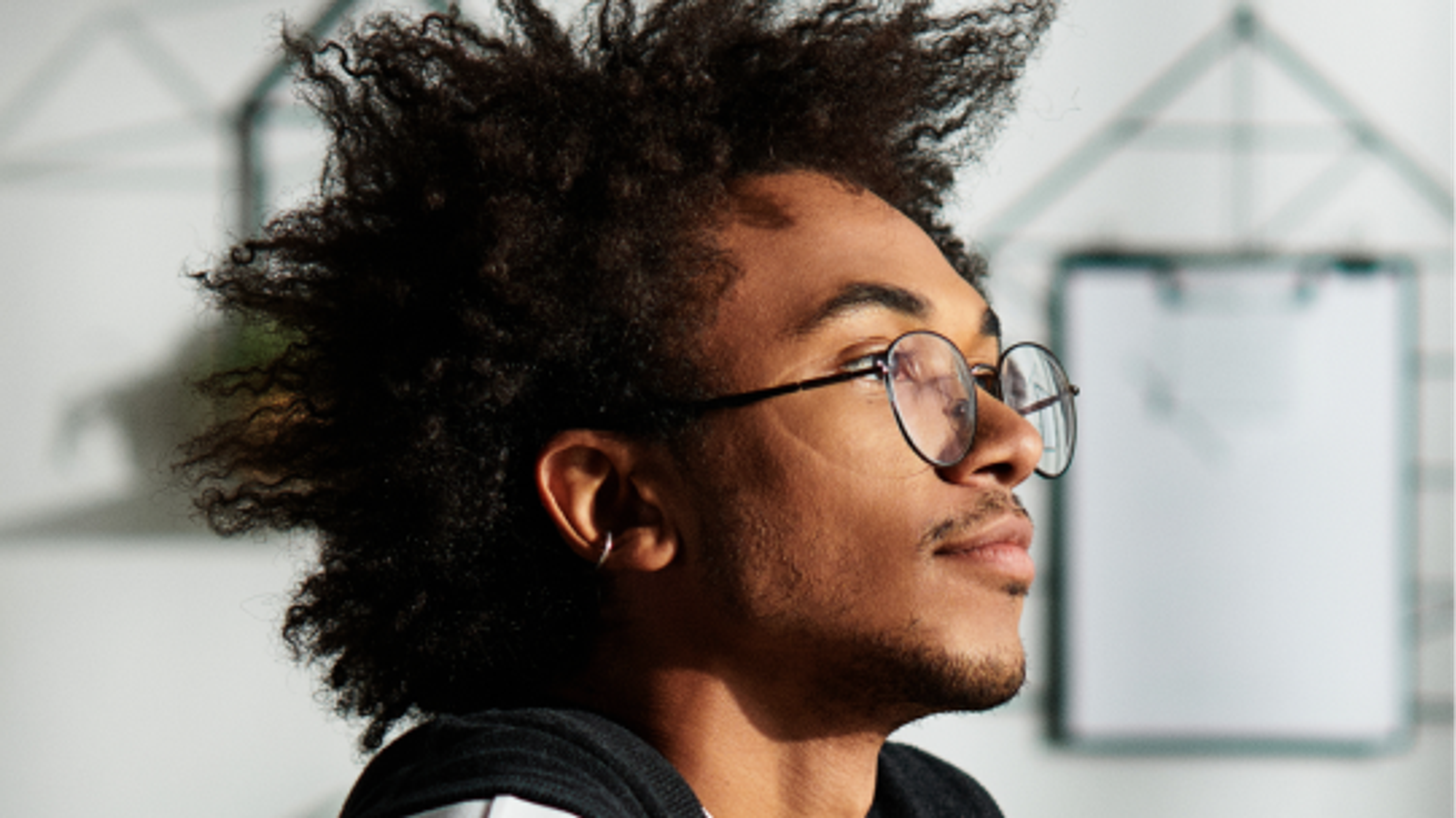 A person with curly hair and glasses looks thoughtfully into the distance, with a blurred background featuring geometric shapes and a clipboard.