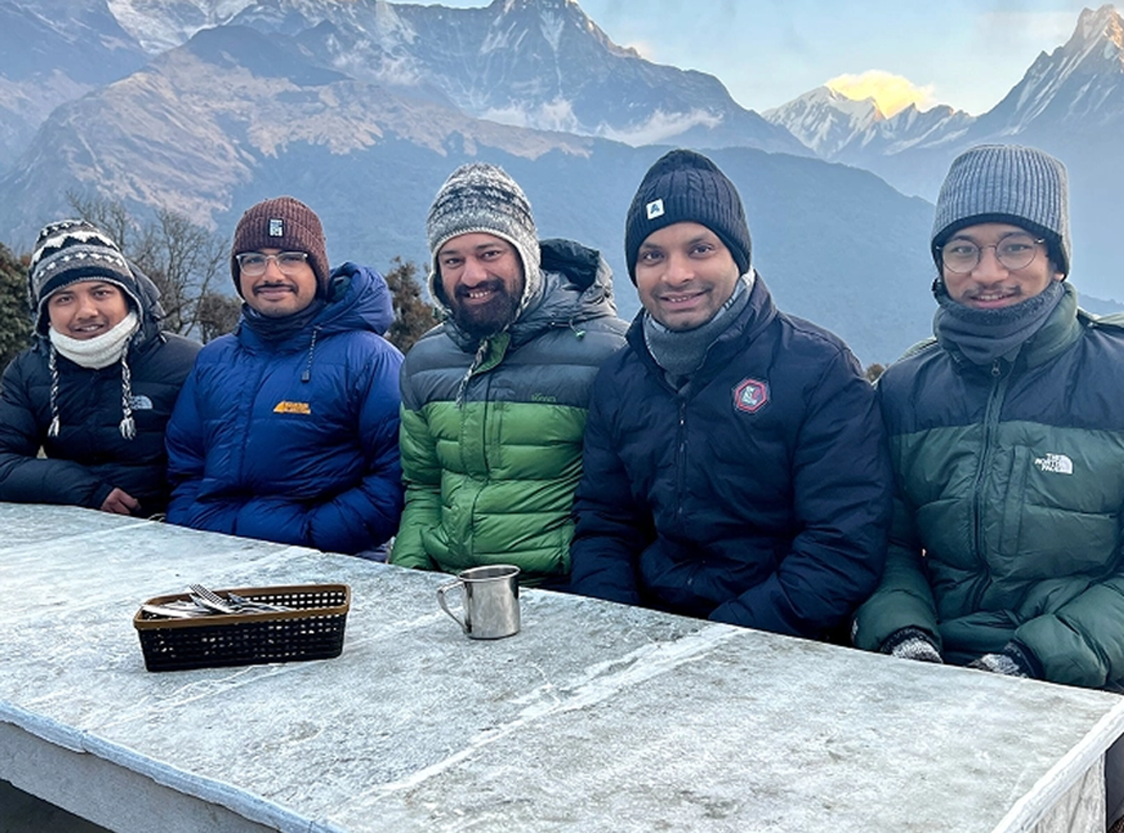 Five people in winter jackets and hats sit at a frosty outdoor table with a metal cup and basket, with snow-covered mountains in the background.
