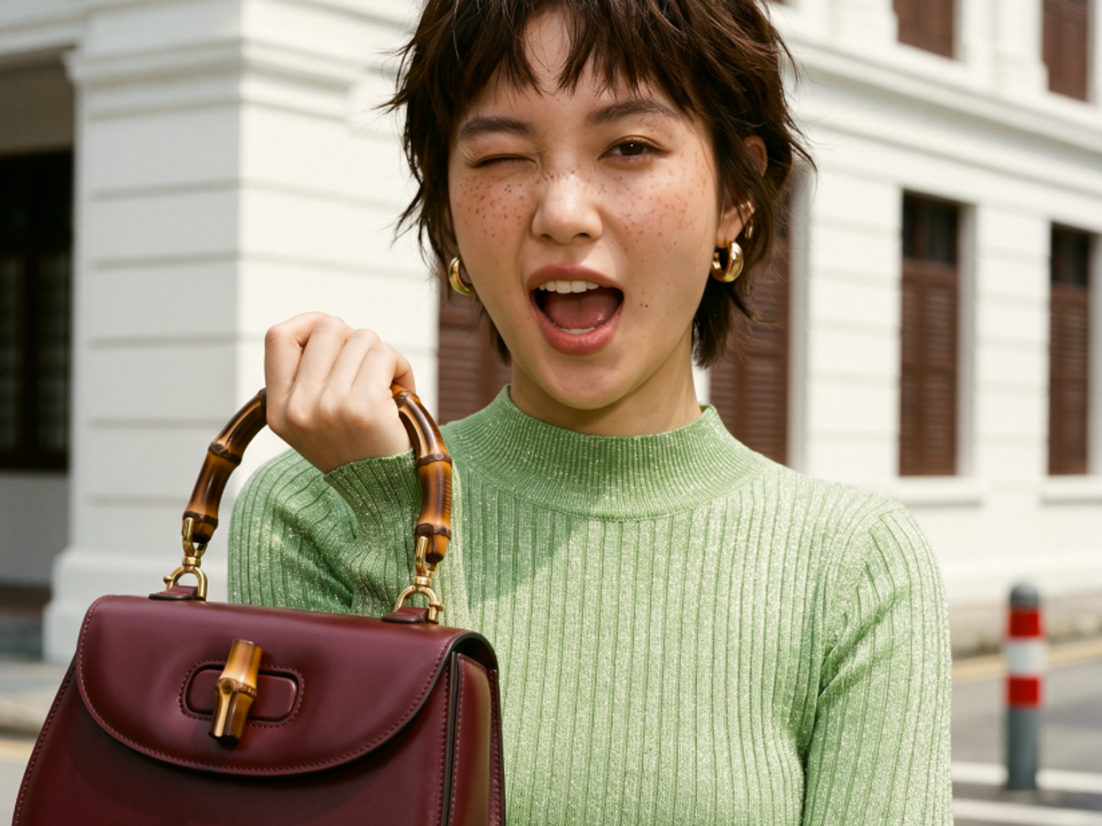 A young woman with short hair and freckles winks while holding a burgundy handbag with a bamboo handle. She is wearing a light green ribbed top and standing outside a white building with brown shutters.