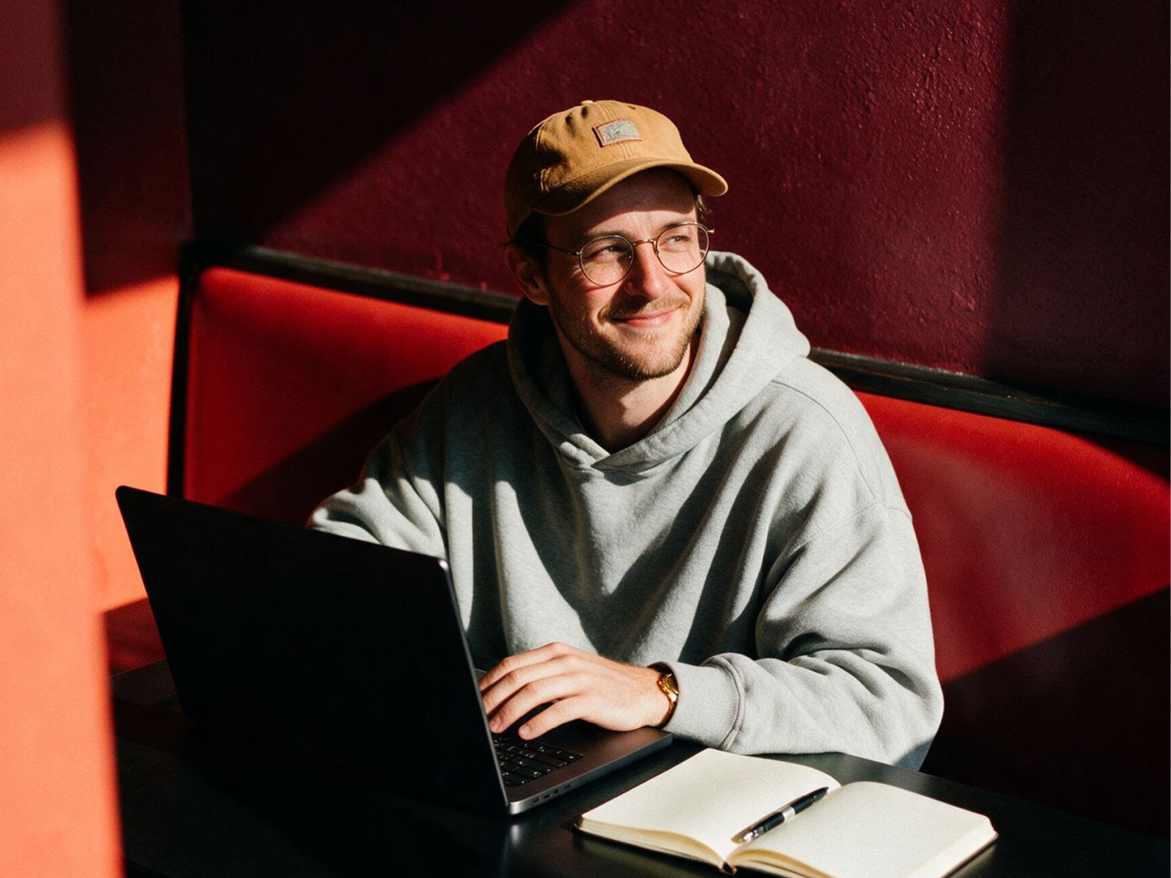 A person wearing glasses, a tan cap, and a gray hoodie sits at a table with a laptop and an open notebook, smiling while looking to the side. Sunlight casts shadows across the red wall and bench behind them.