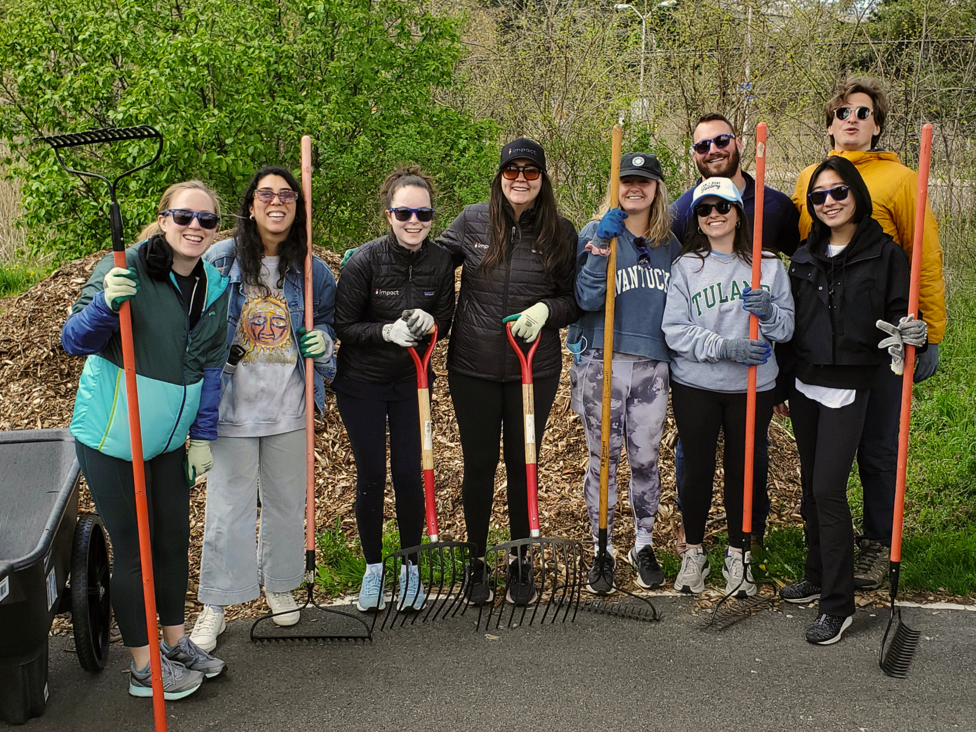 Nine people stand outdoors holding rakes and smiling at the camera, wearing gloves and sunglasses, with a large pile of mulch and green foliage in the background.