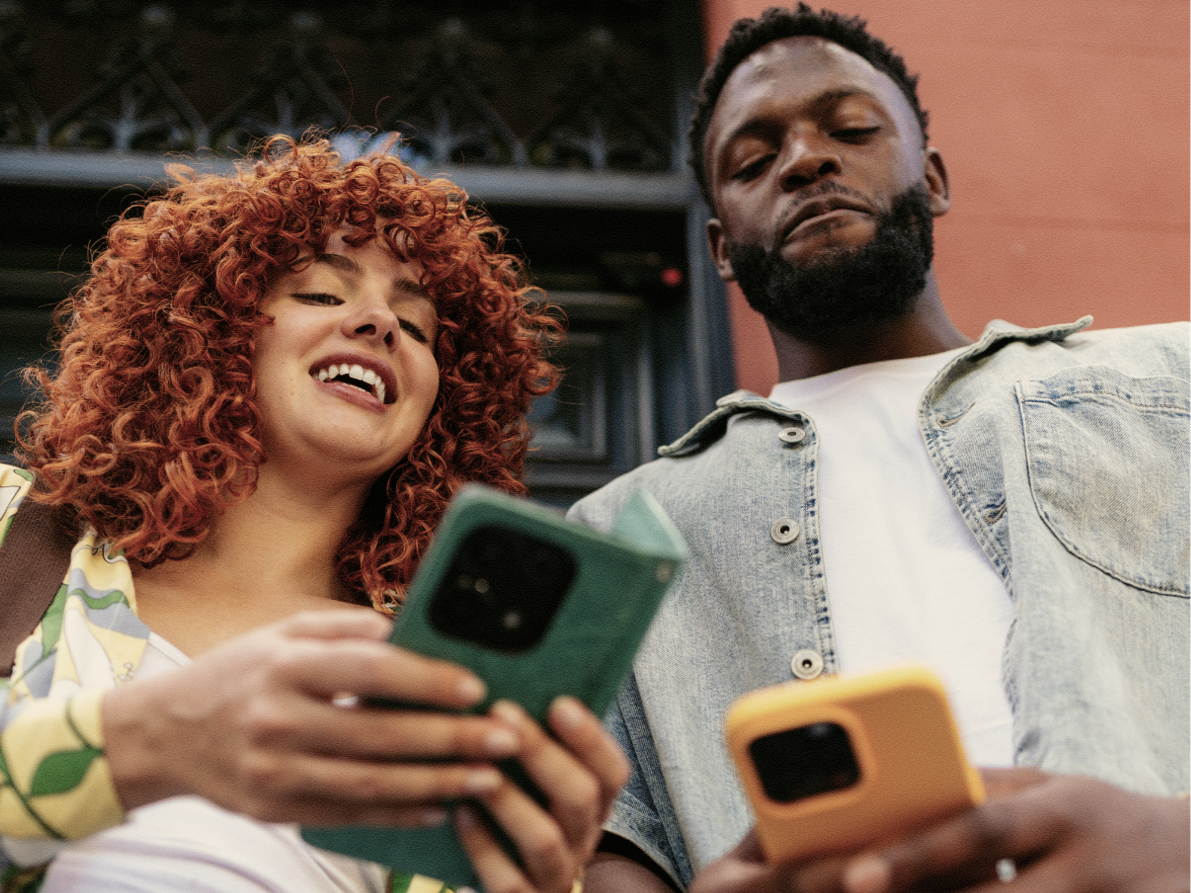 Two people standing outdoors, smiling and looking down at their smartphones. One has curly red hair and the other has short dark hair and a beard. Both appear engaged with their devices.