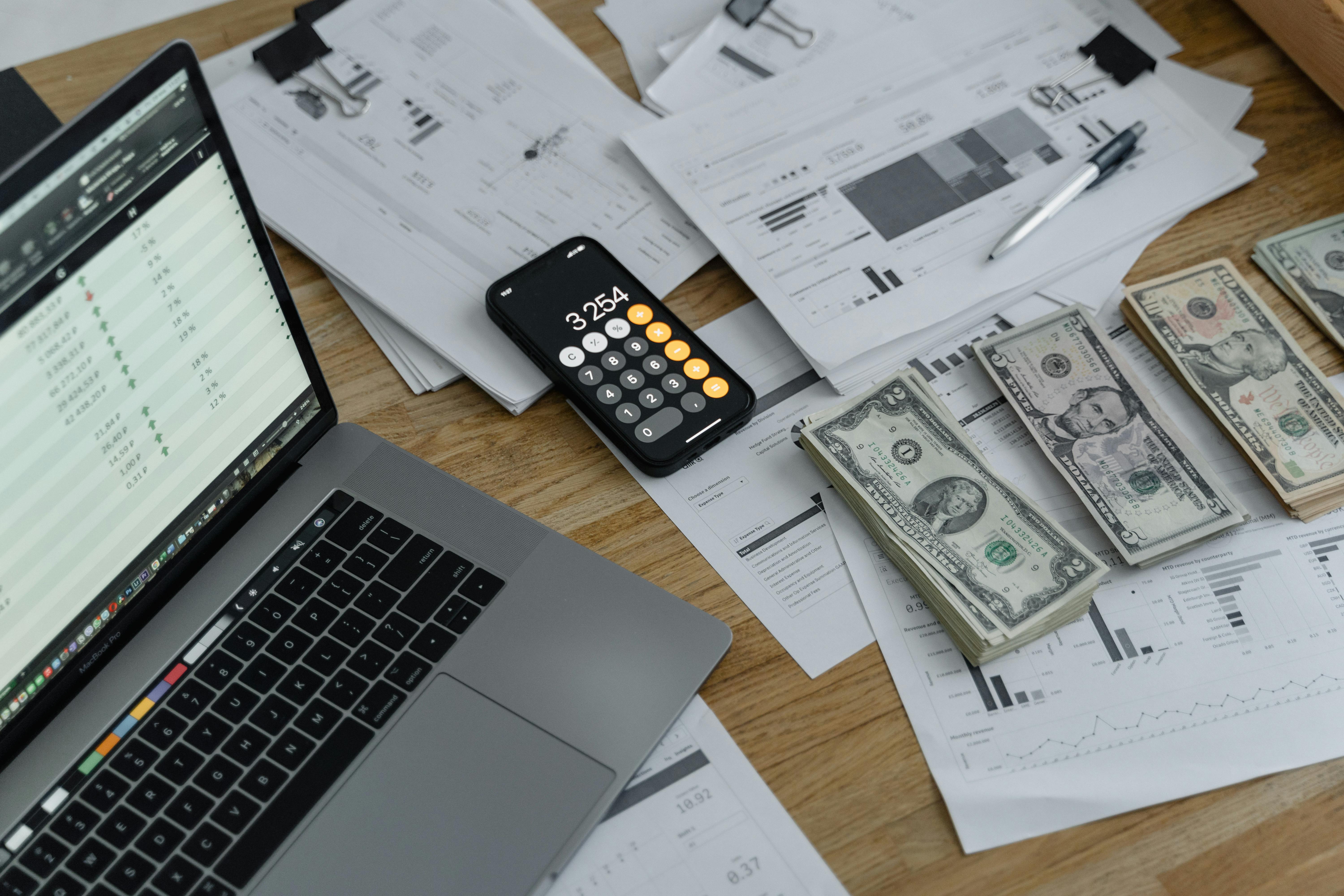 desk with laptop, stacks of money and iphone showing calculator app