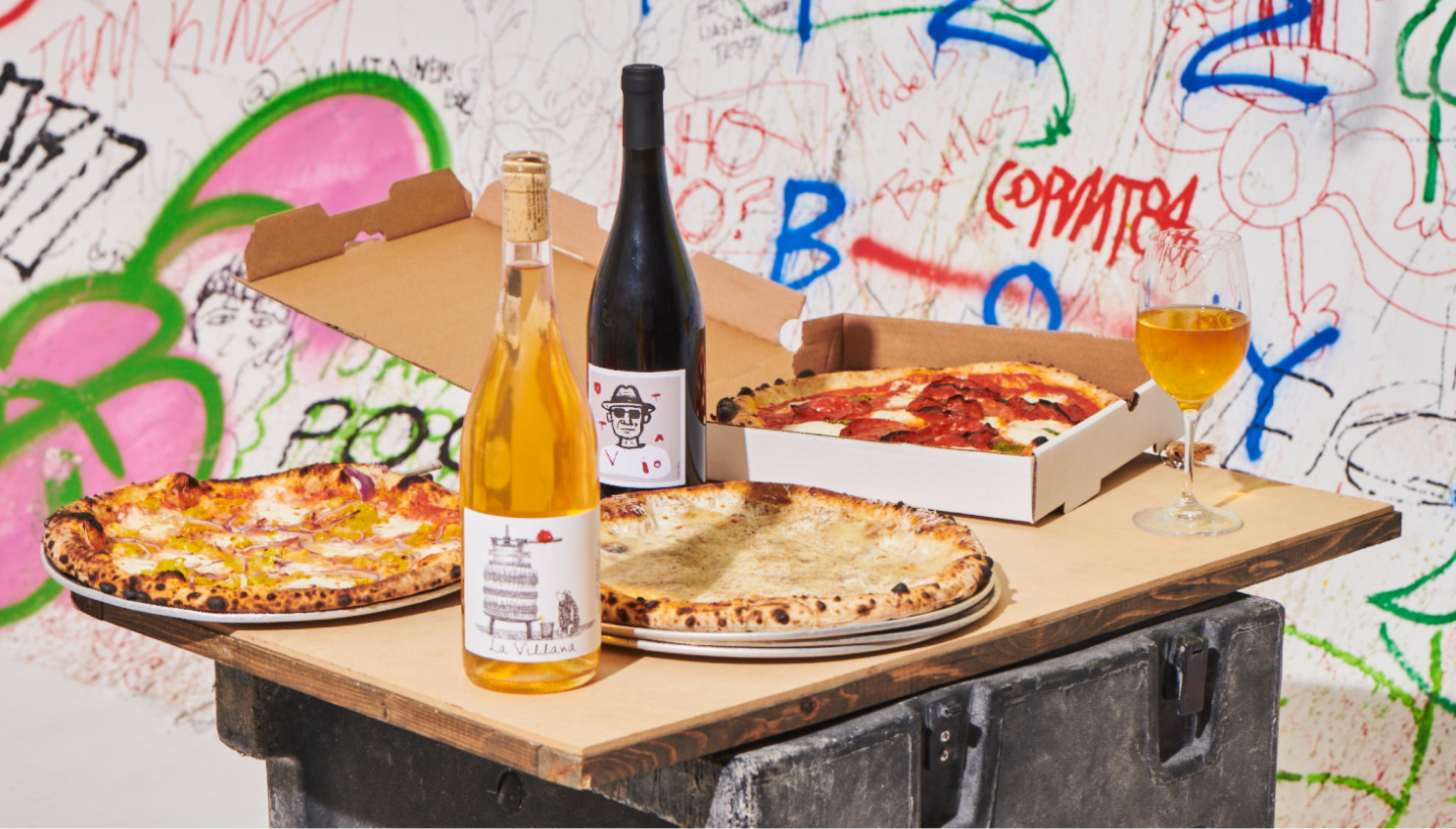 photograph of spread of pizza and wine set against a wall of graffiti