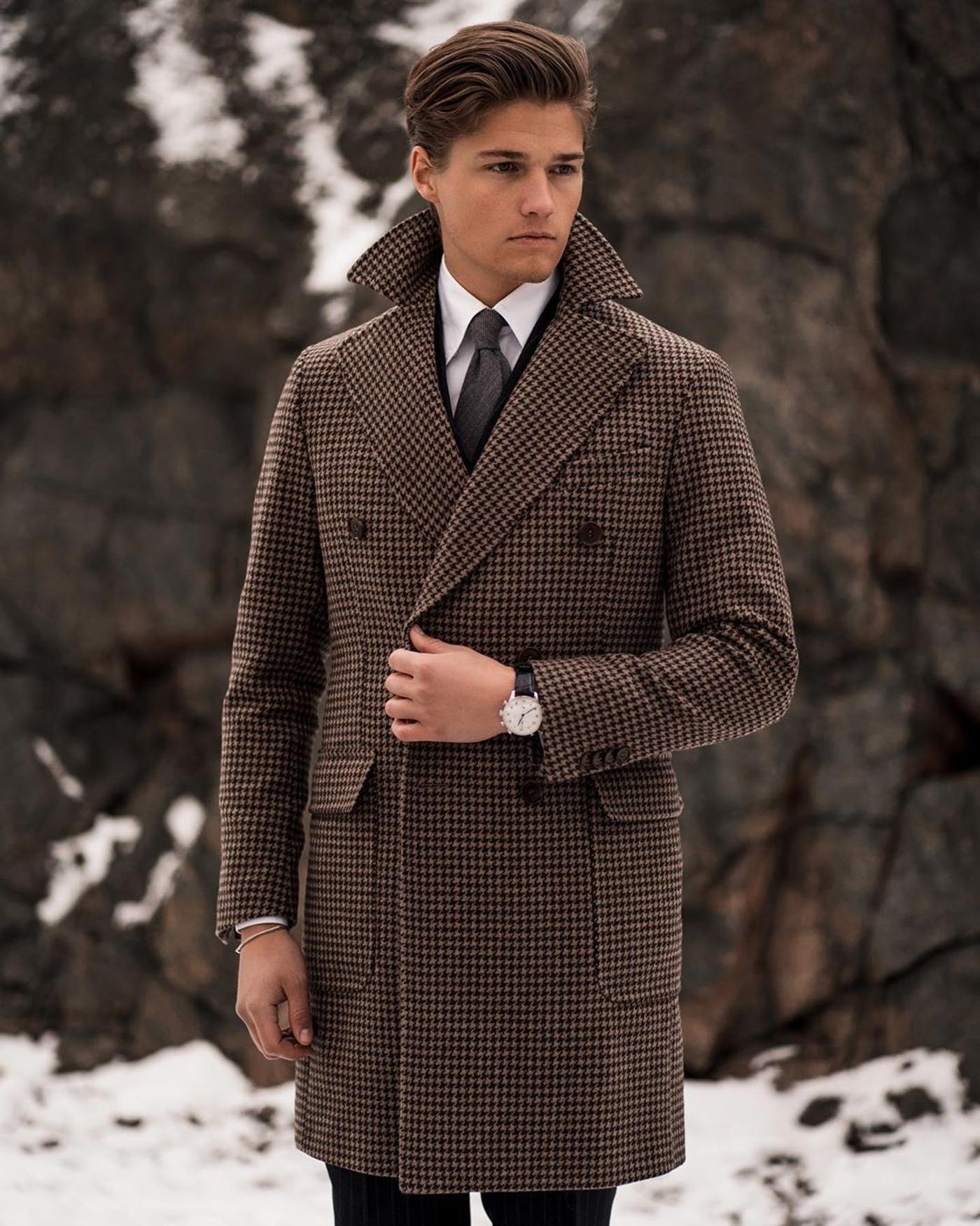 A stylish young man in a brown houndstooth double-breasted overcoat and white shirt stands in a snowy, rocky setting.