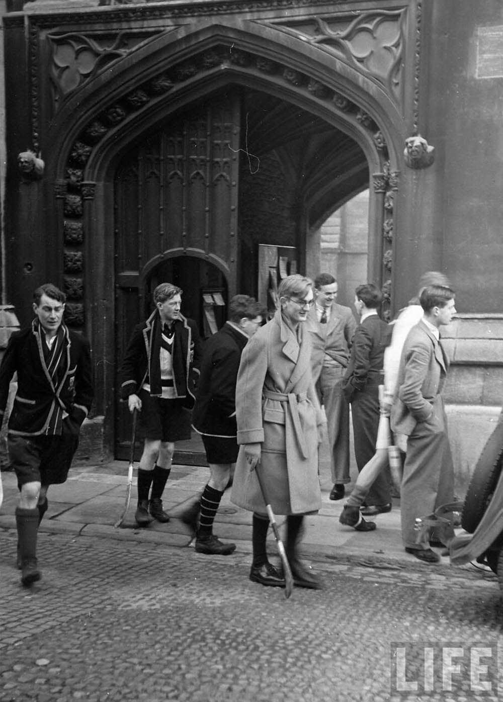 Students in varying formal and sporty attire walk on a cobblestone path in front of a large gothic archway, some carrying sticks.