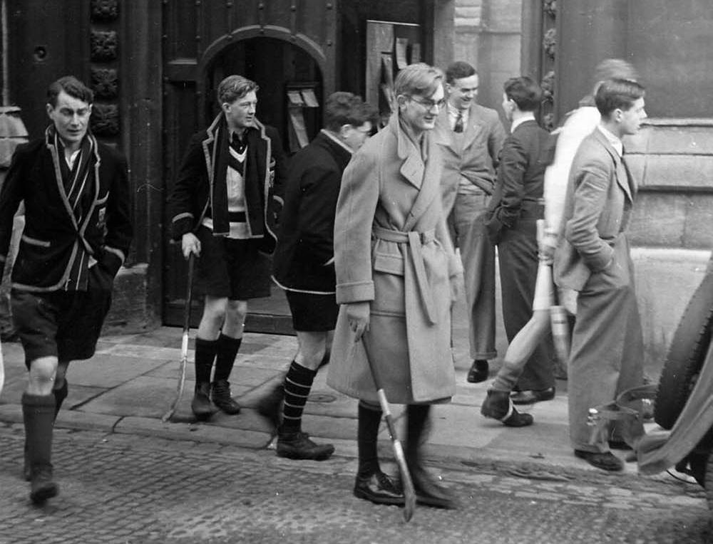 Students in varying formal and sporty attire walk on a cobblestone path in front of a large gothic archway, some carrying sticks.