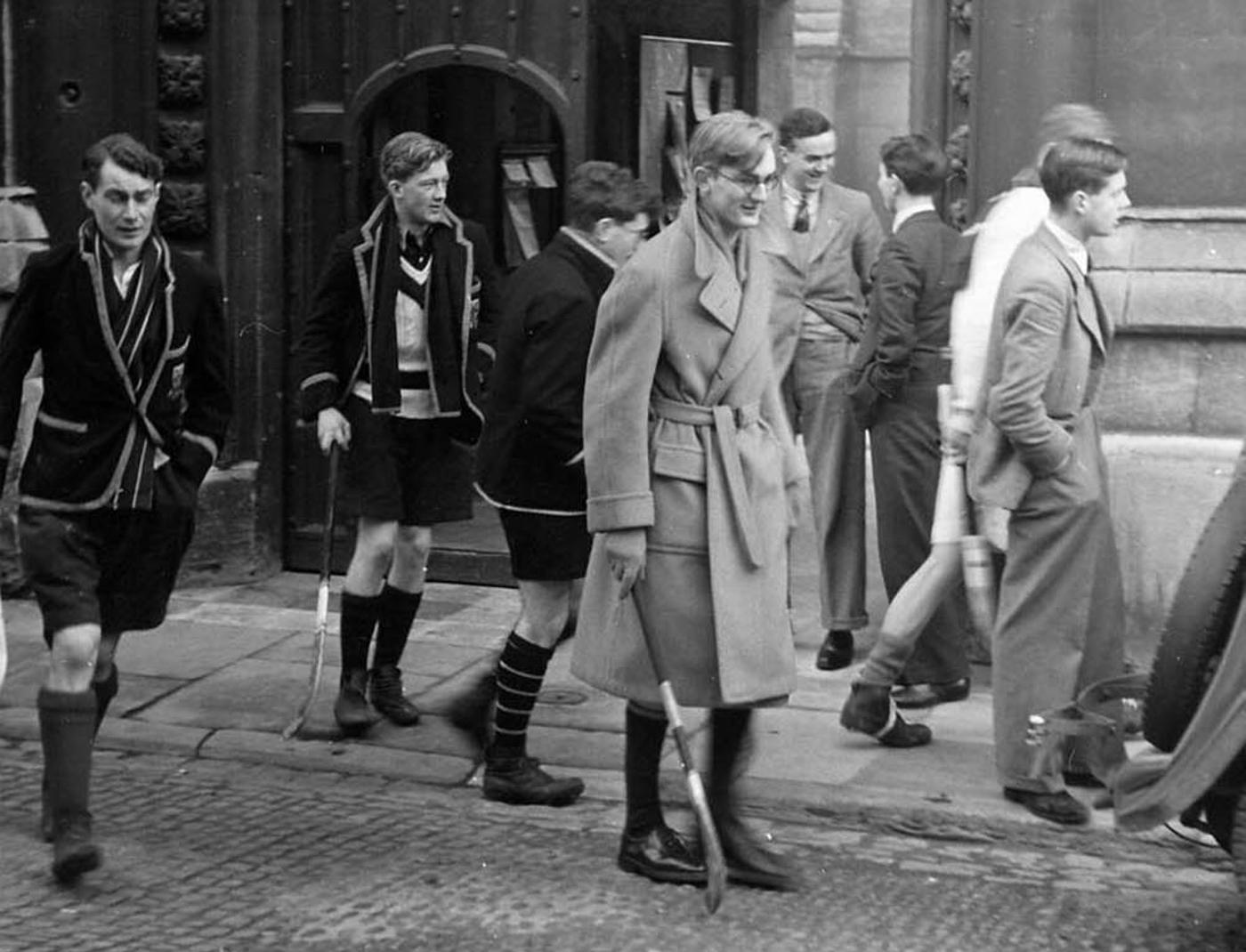 Students in varying formal and sporty attire walk on a cobblestone path in front of a large gothic archway, some carrying sticks.