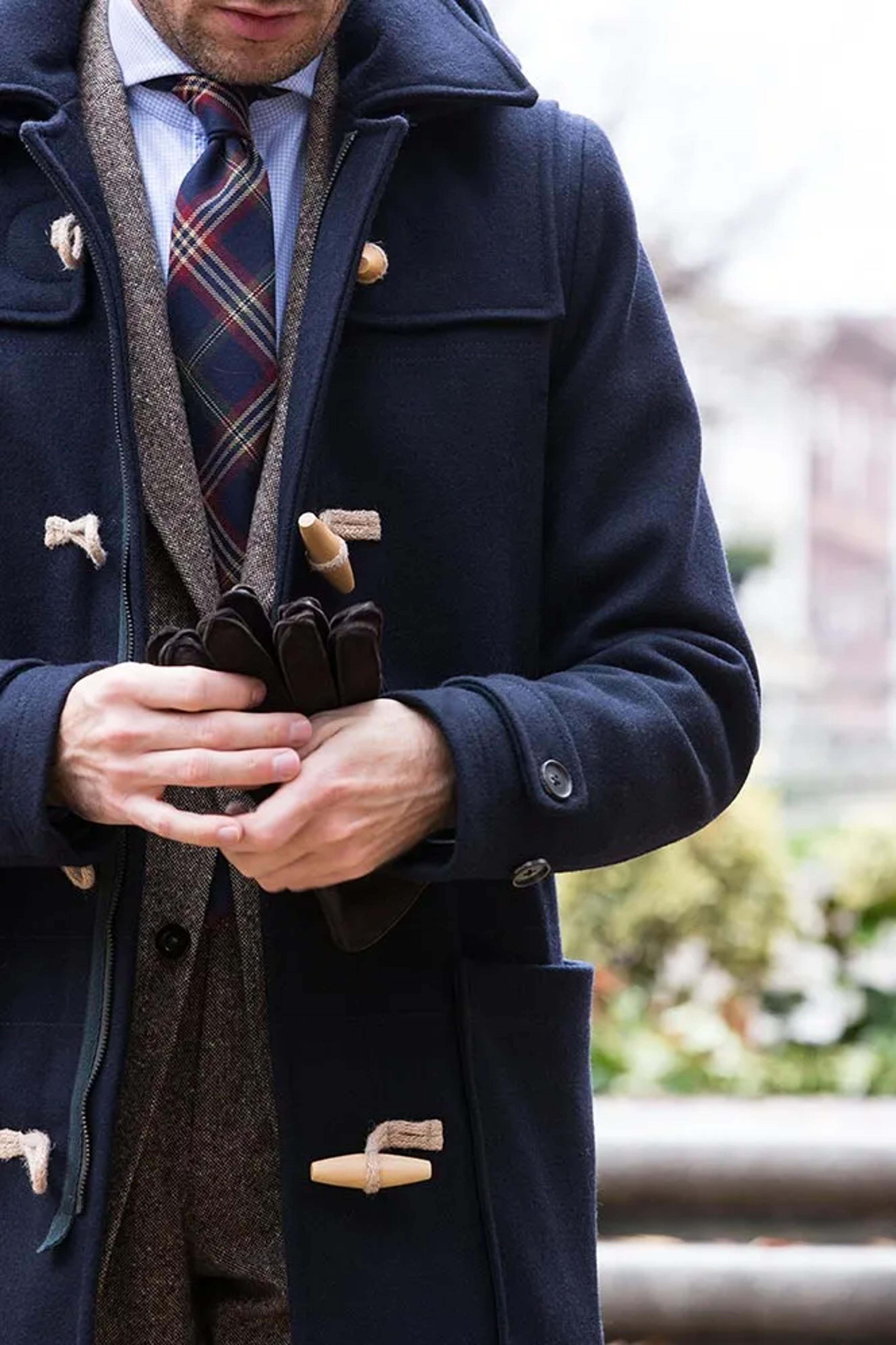 A man in a navy duffle coat, plaid tie, and tweed jacket holds brown gloves.