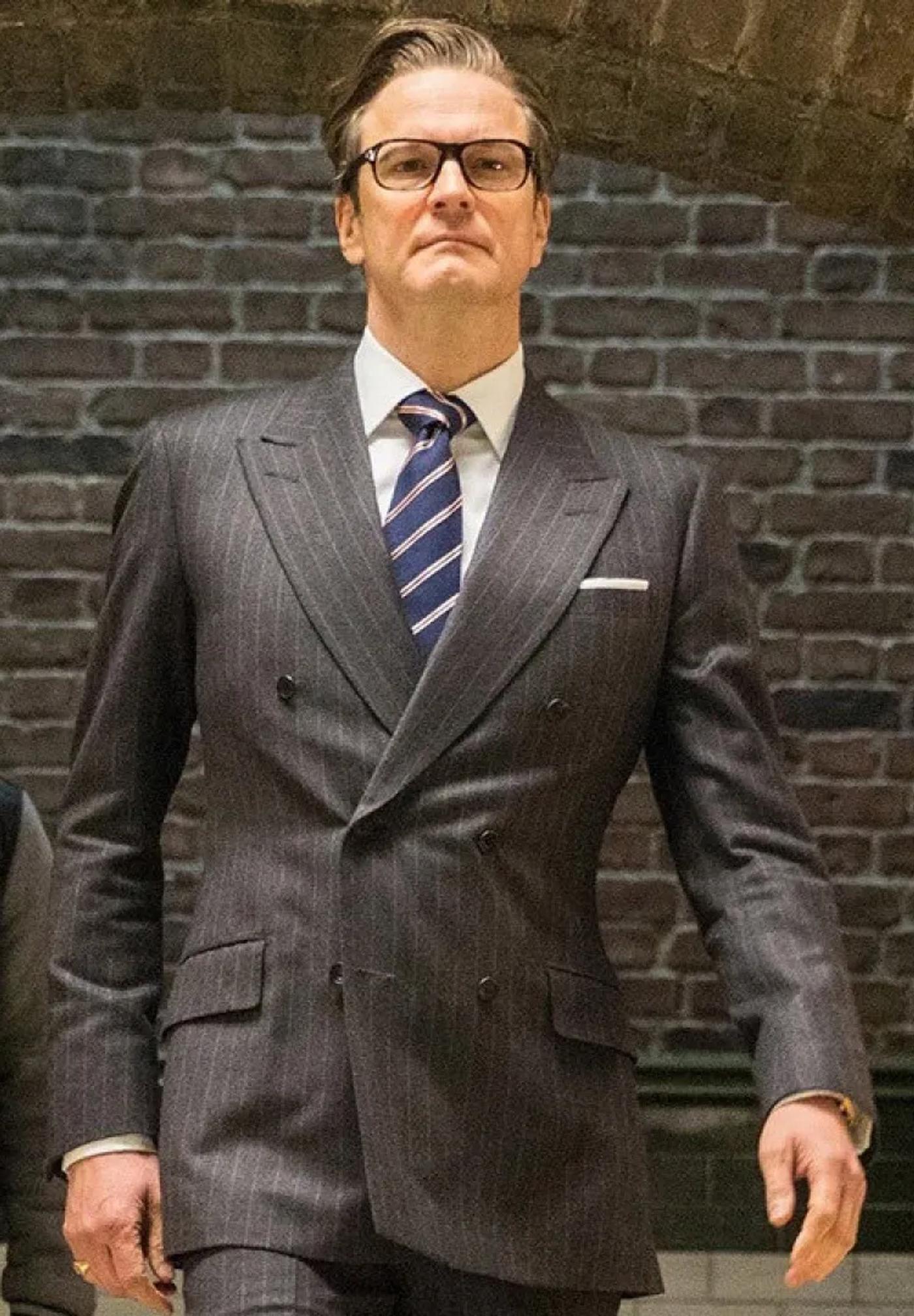 Colin Firth as Harry Hart, wearing a dark pinstripe double-breasted suit, white shirt, striped tie, and glasses, looking directly at the viewer against a brick wall.