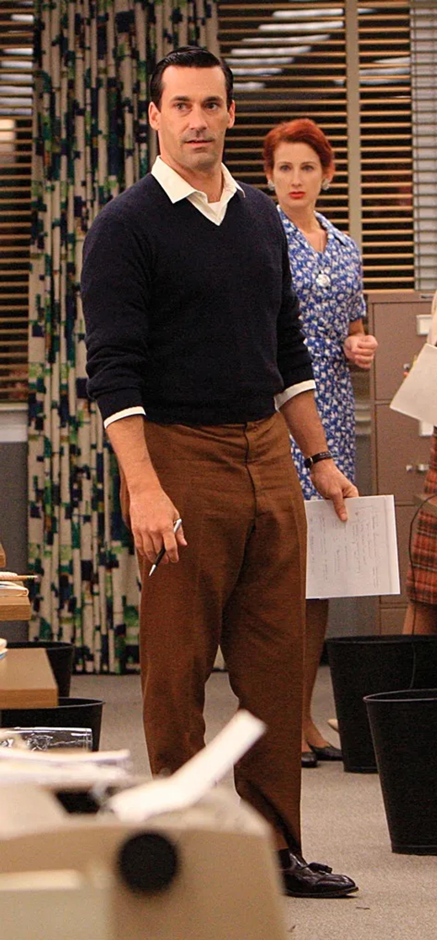 Jon Hamm as Don Draper in a navy sweater and brown pants, holding papers and a pen, with a woman in a blue dress behind him in an office.