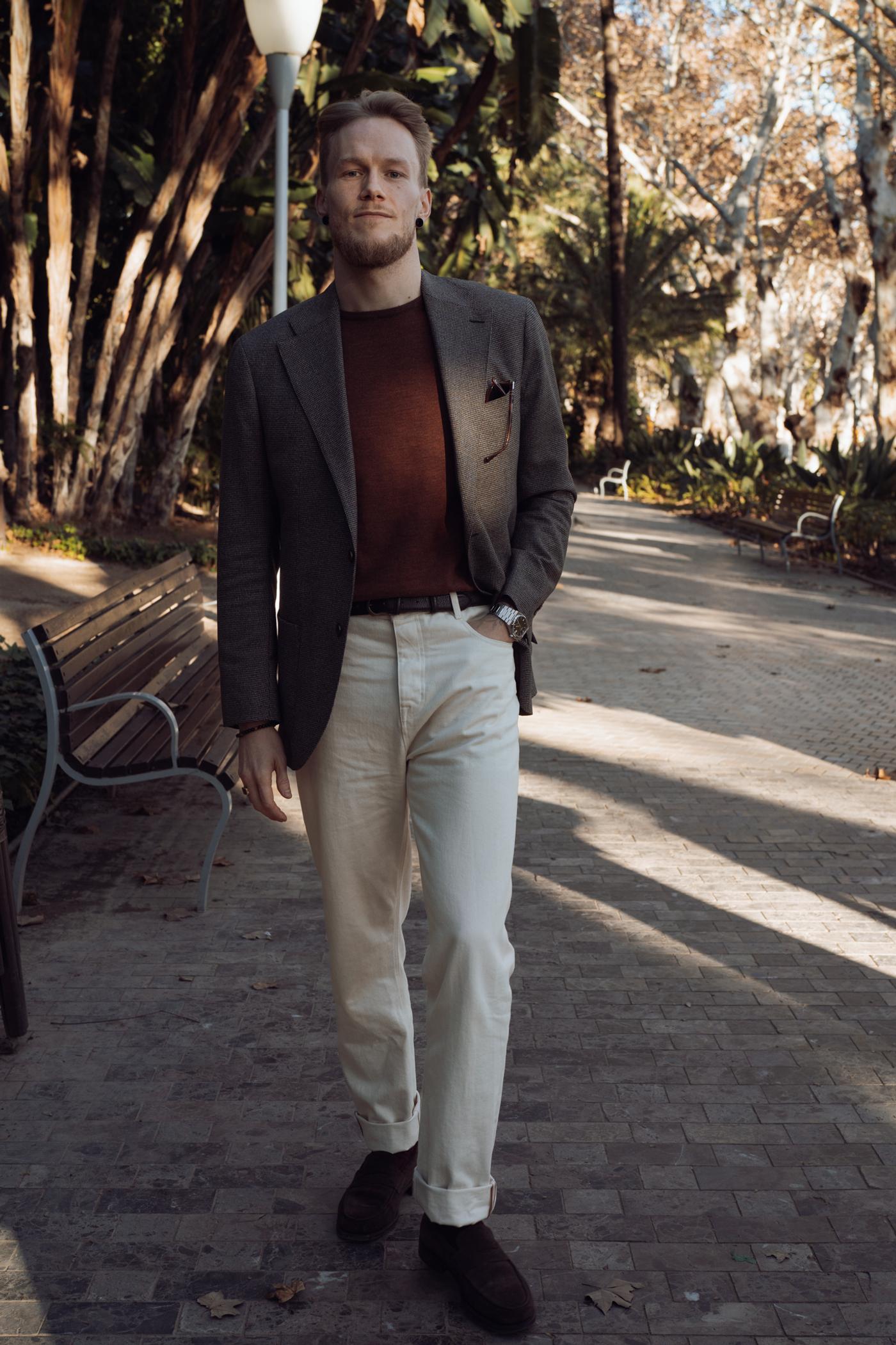 A man wearing a brown blazer, rust sweater, and cream pants walks on a sunlit paved path in a park.