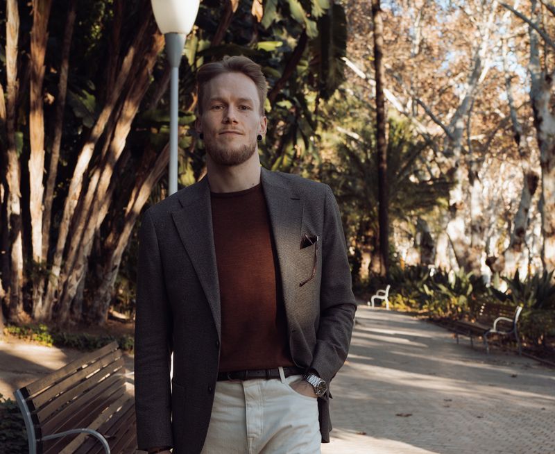 A man with a beard and blond hair stands in a sunny park, wearing a grey blazer, rust-colored shirt, and light pants.