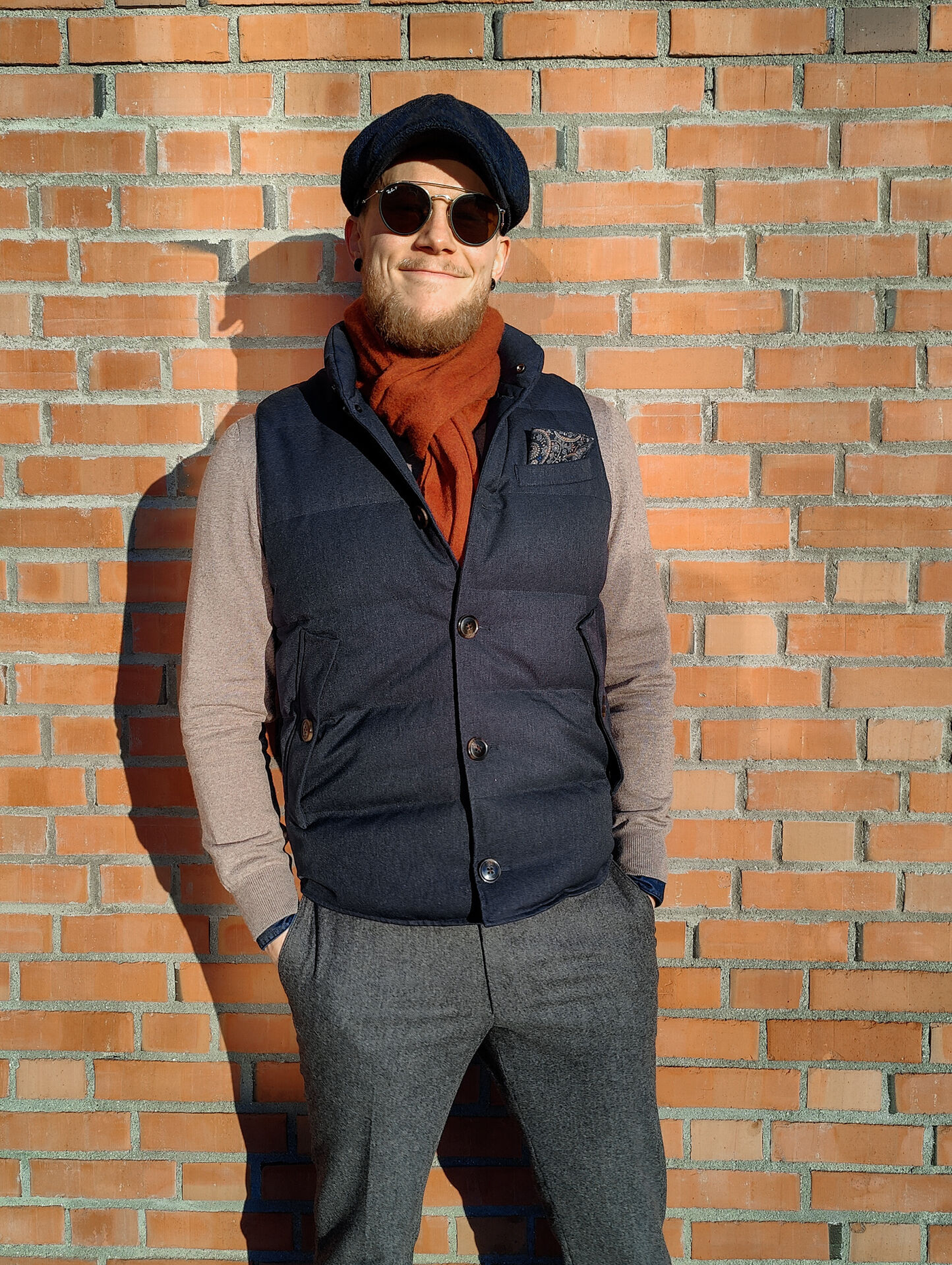 Kristoffer wearing gray flanner trousers with a sand cashmere sweater, a navy vest, orange scarf and blue bakerboy hat