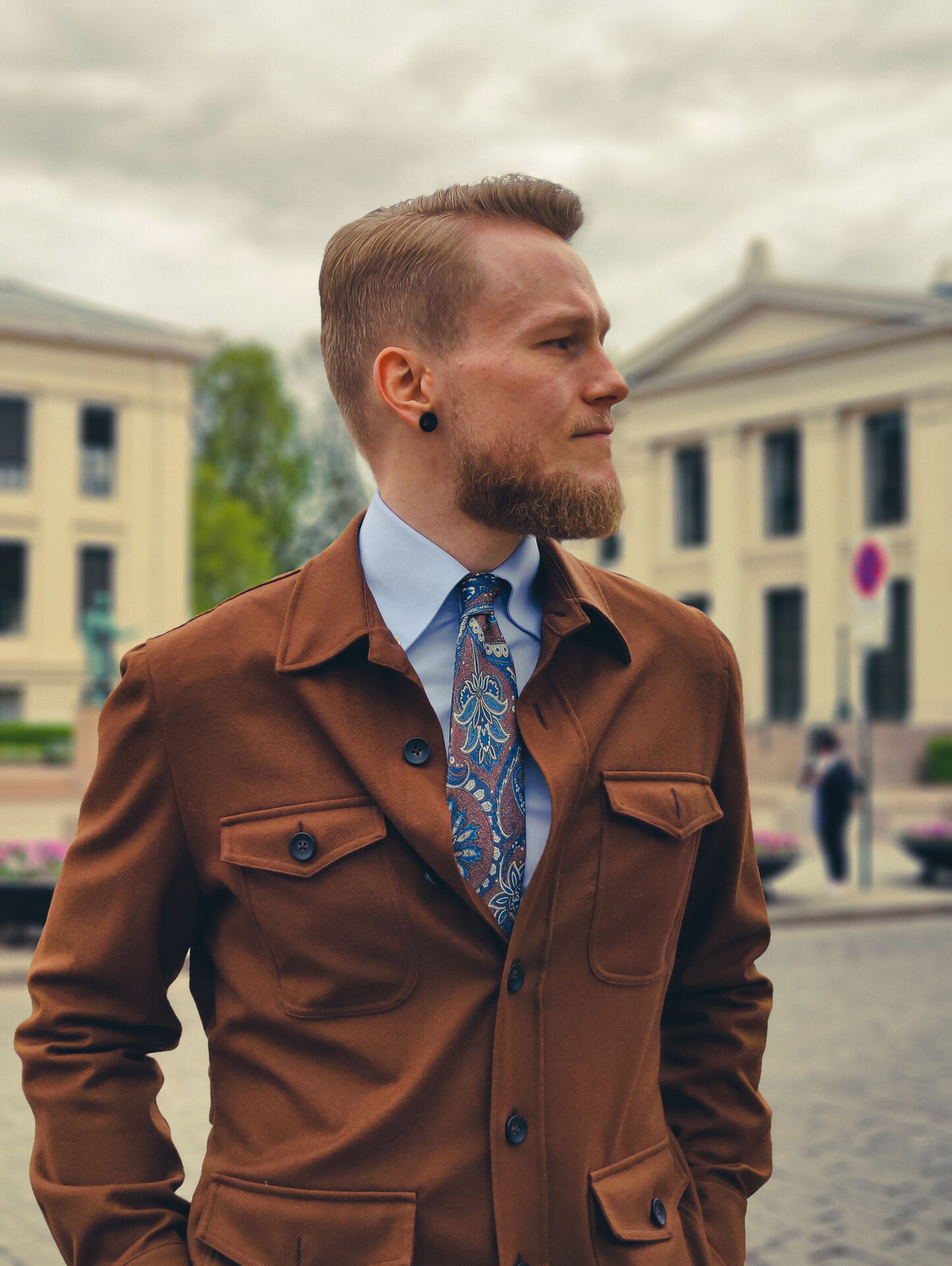 A bearded man in a brown safari jacket and patterned tie looks right in an urban setting.