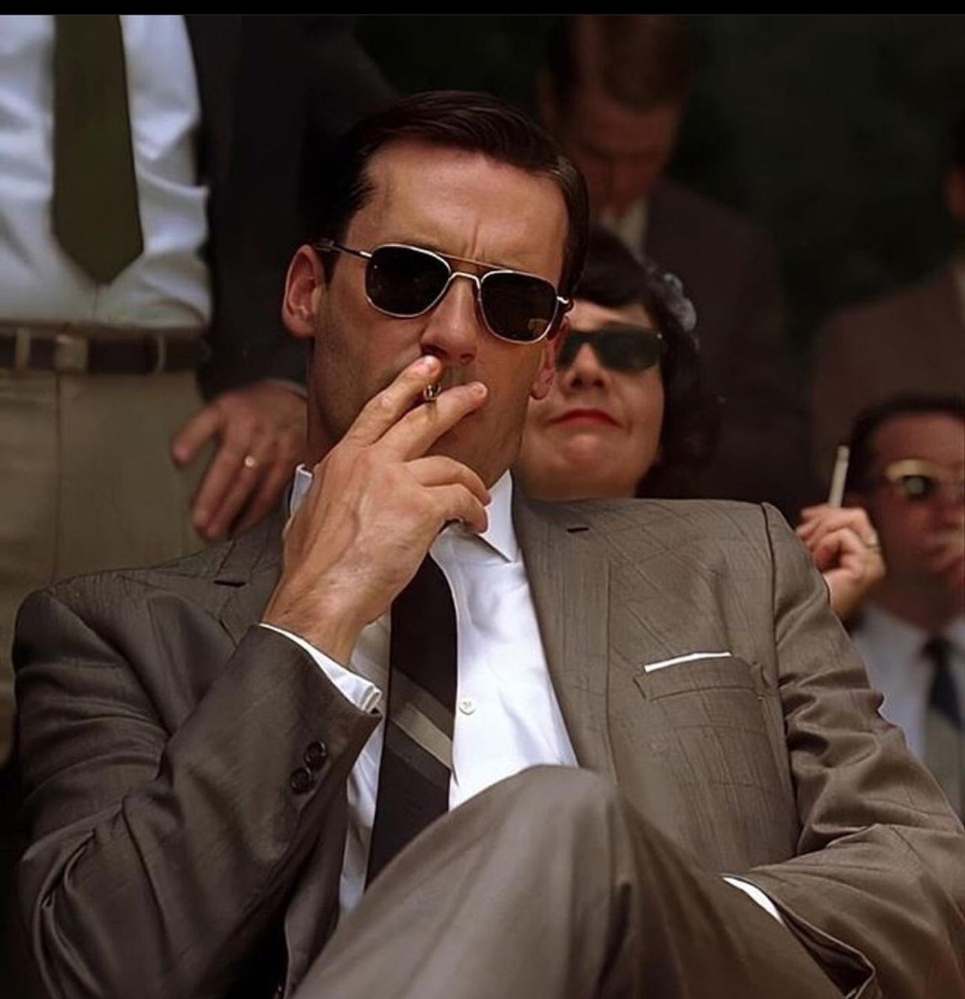 Don Draper in a grey suit and aviator sunglasses smokes a cigarette.
