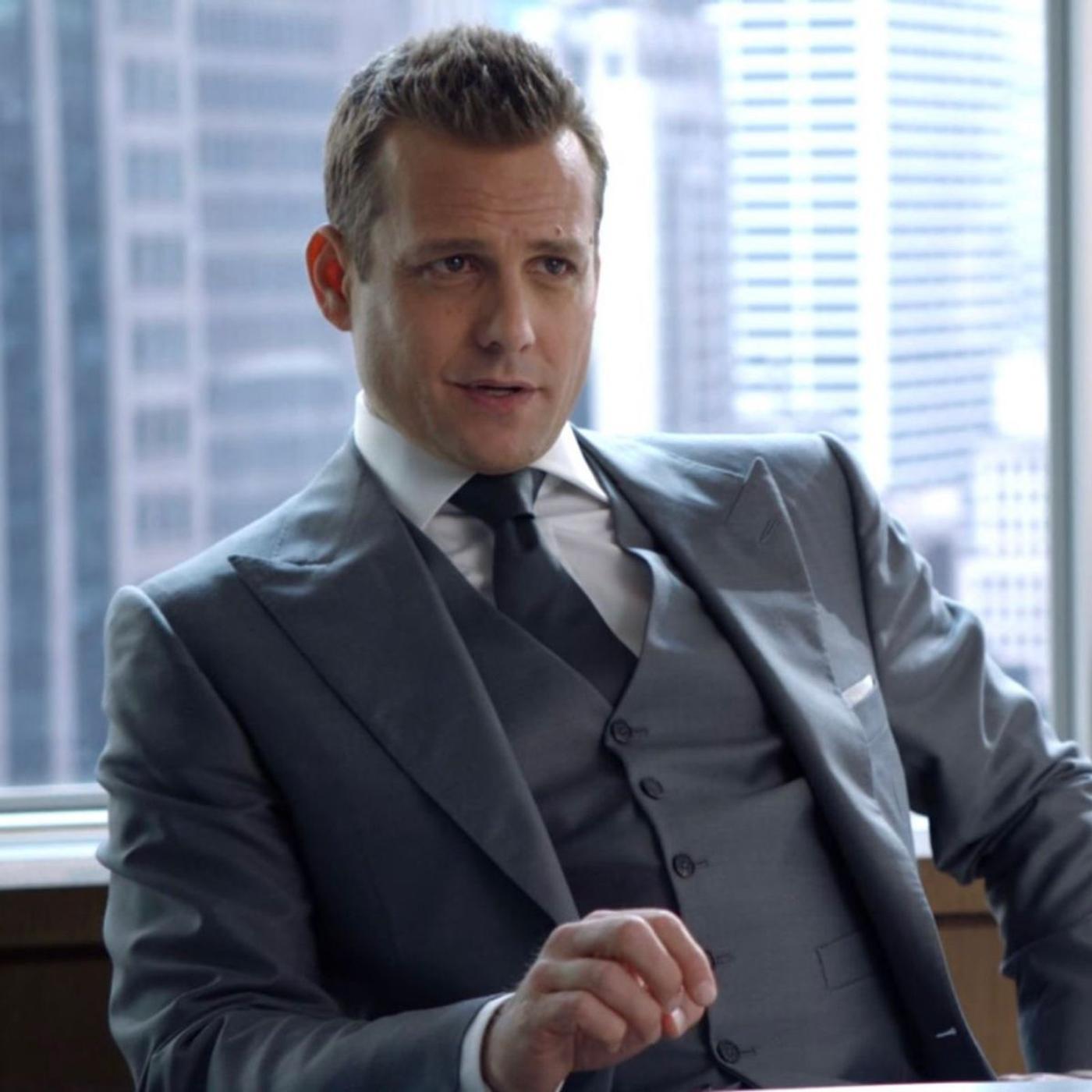 A man in a grey three-piece suit and tie, sitting in an office with a city view.