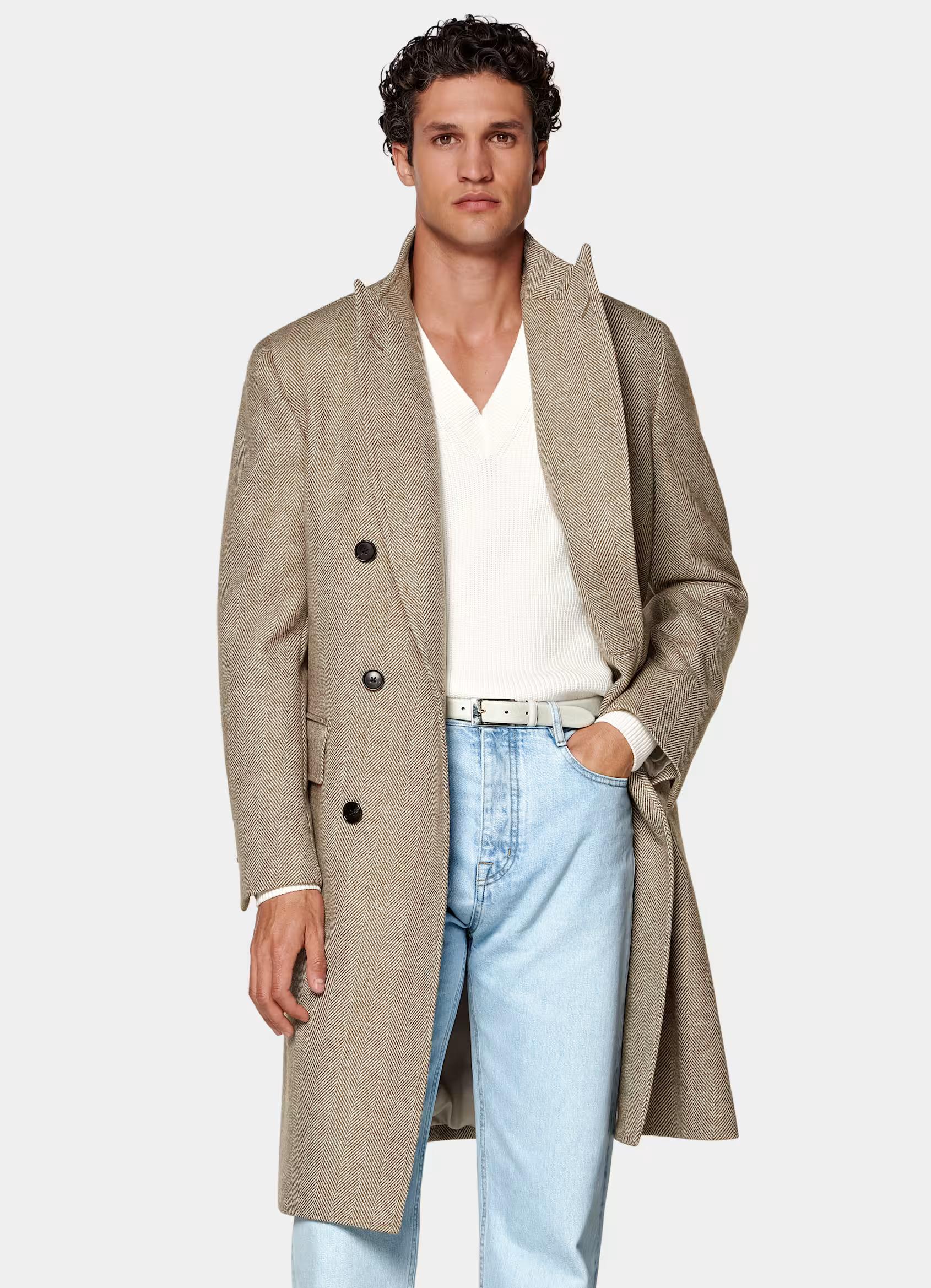 A man in a tan double-breasted coat, white V-neck sweater, and light wash jeans.
