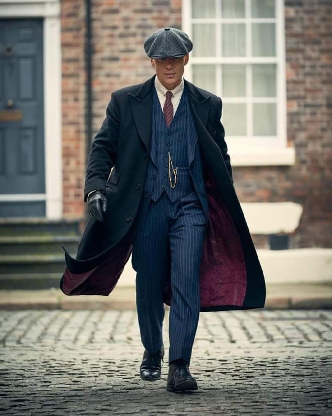 A man in a dark overcoat, pinstripe suit, and flat cap walks on a cobblestone street.
