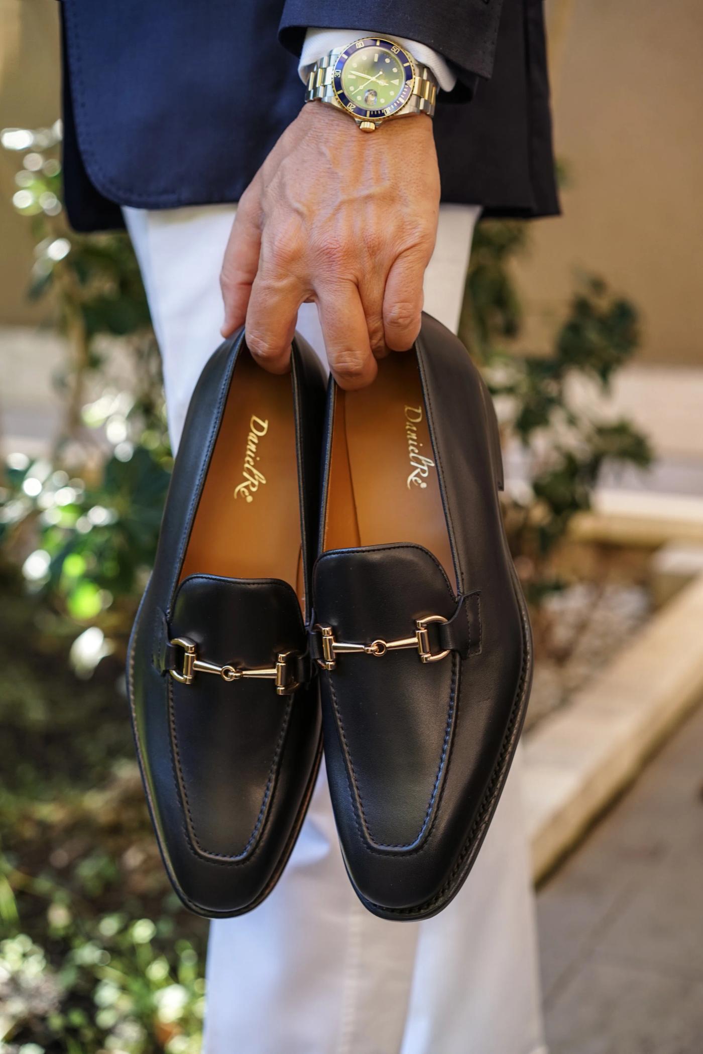 A person holds a pair of black leather loafers with gold horsebit details.
