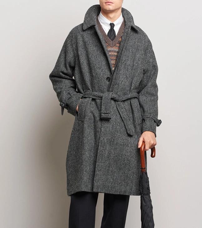 Man in a belted grey herringbone tweed coat, dark trousers, and black shoes, holding a closed black umbrella.