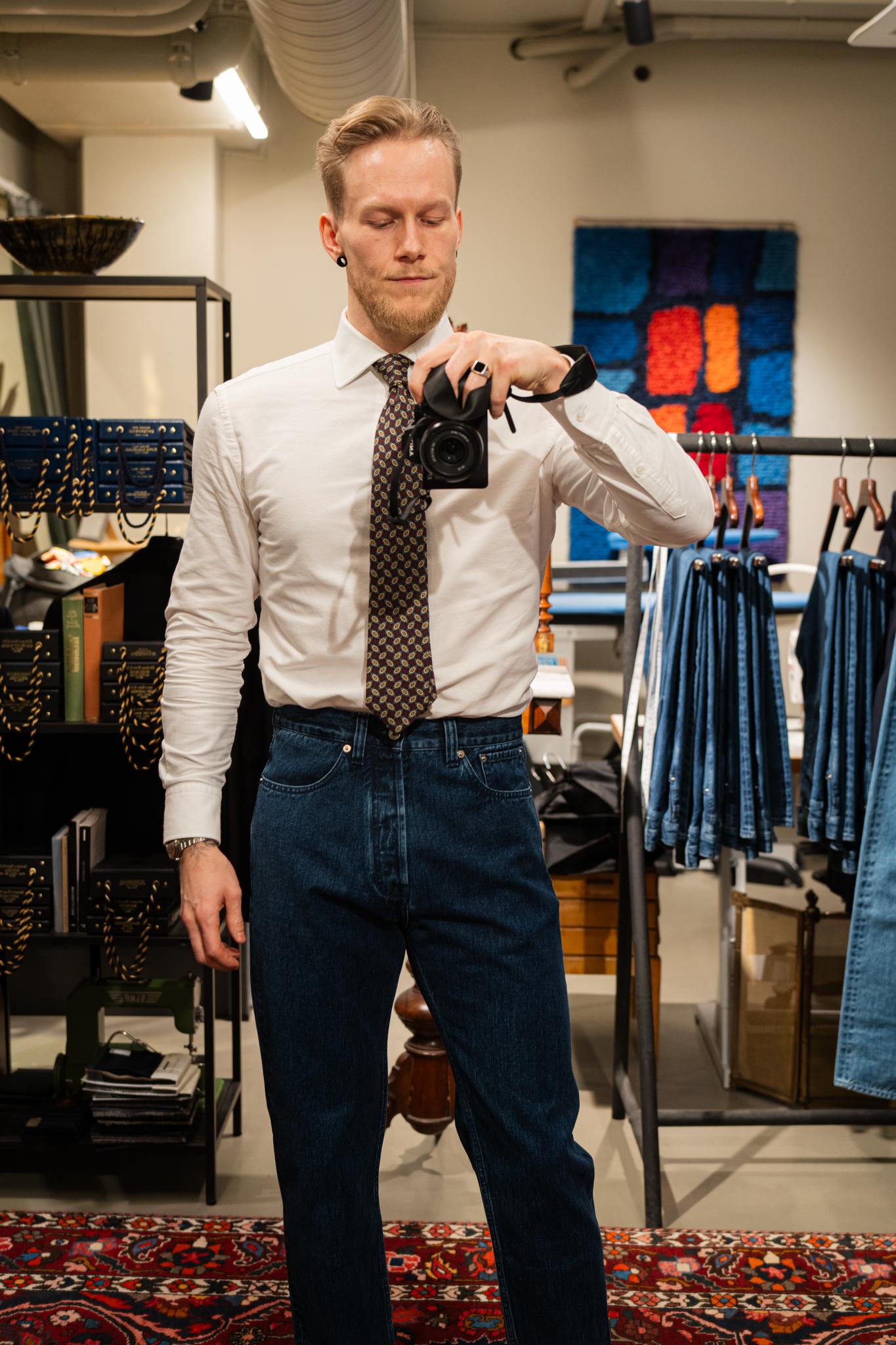 A man in a white shirt, patterned tie, and dark jeans takes a mirror selfie with a camera in a store.