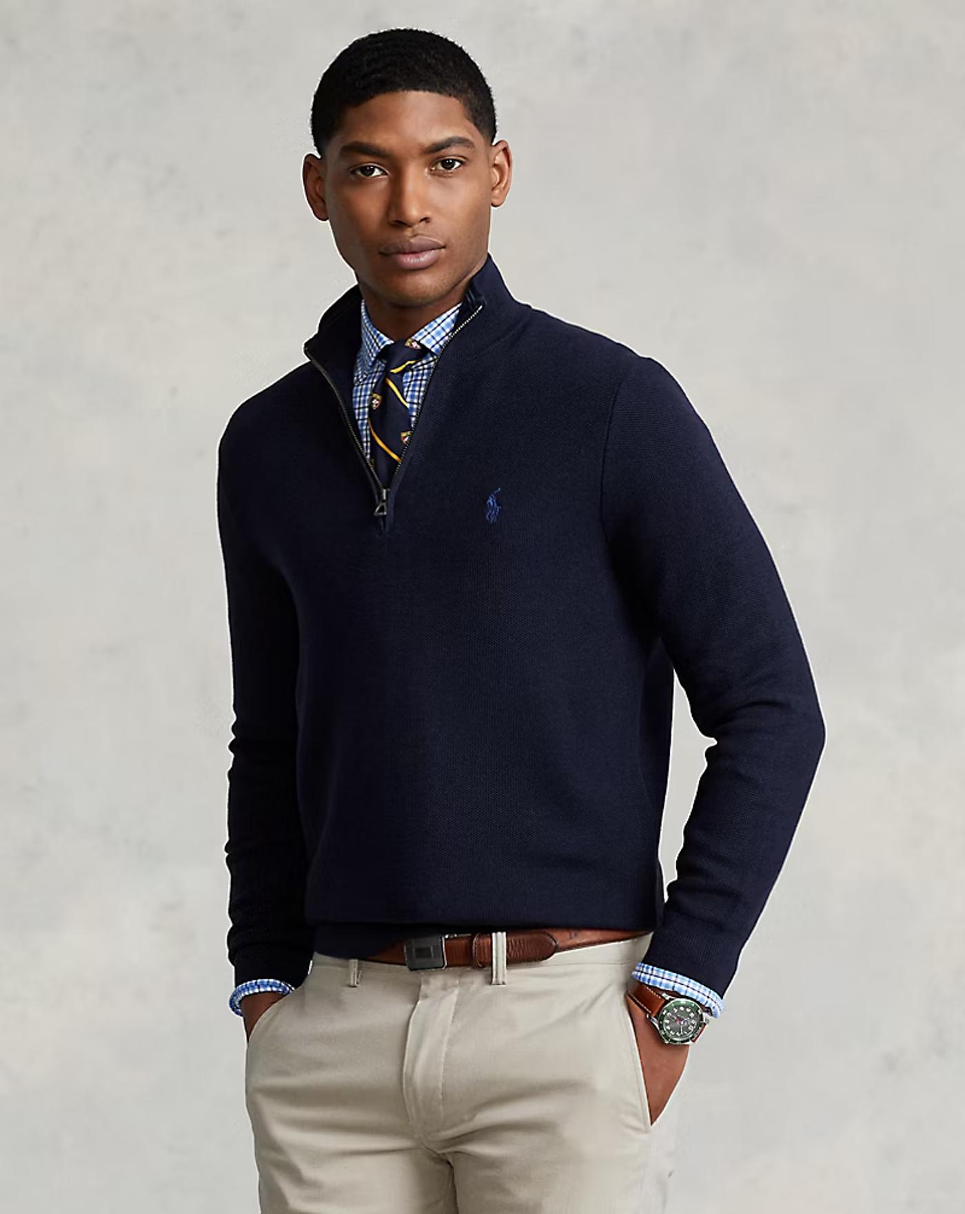 A man in a navy Ralph Lauren quarter-zip sweater over a blue checkered shirt and striped tie, with khaki pants, looks at the camera.