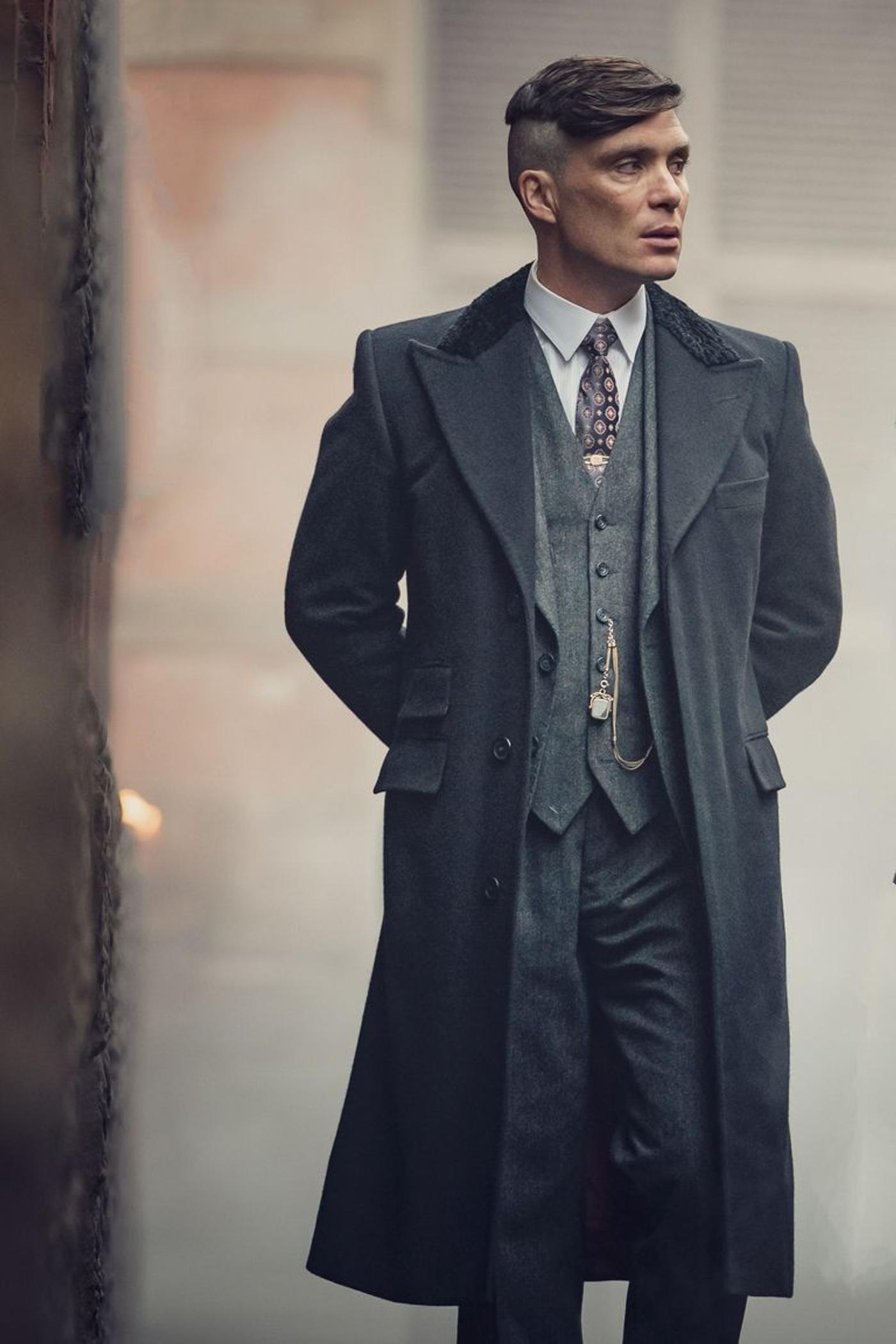Tommy Shelby (Cillian Murphy) in a dark overcoat and suit, hands clasped behind his back, looking right in a smoky setting.