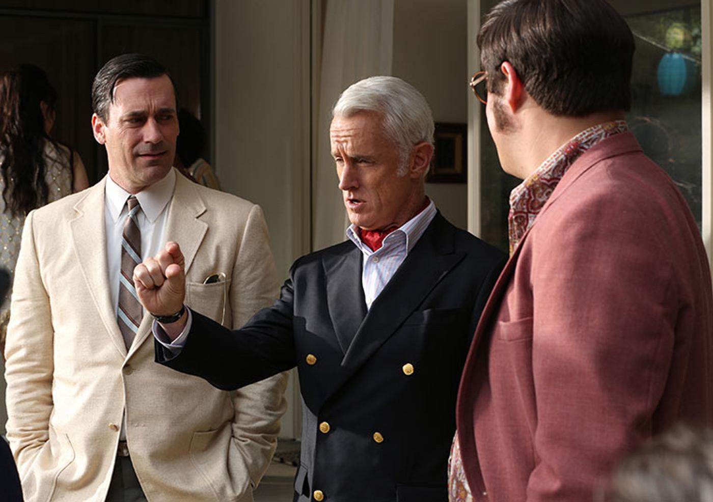 Three men in retro clothing; a man in a light suit watches a white-haired man in a dark jacket and red ascot who is gesturing, facing a man in a pink jacket seen from behind.