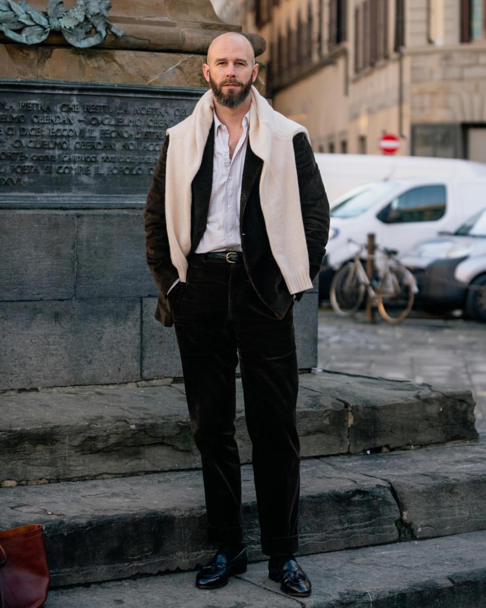 a bald man wearing a suit and scarf stands in front of a statue