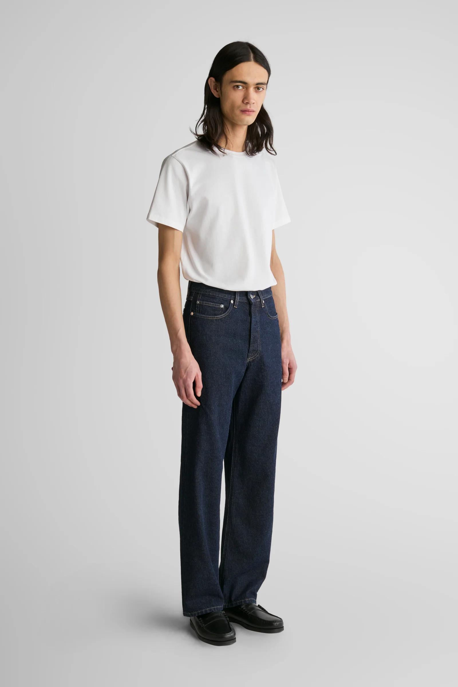 A man with long dark hair wearing a white t-shirt, dark blue jeans, and black loafers.