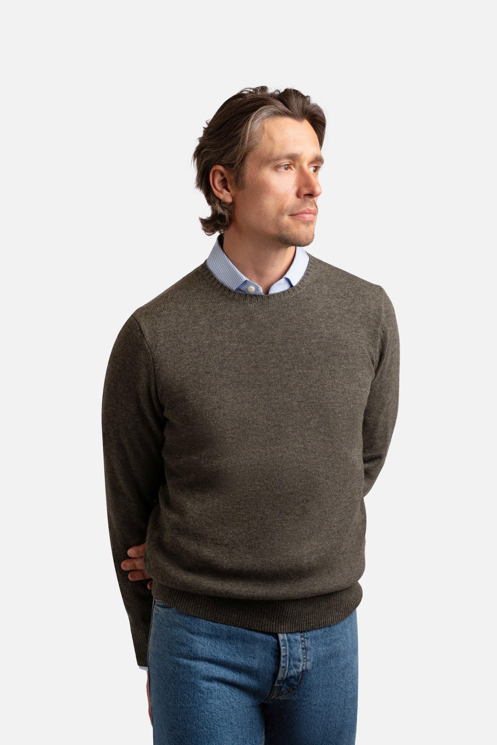 A man wearing an olive green crewneck sweater over a light blue collared shirt and blue jeans, looking to the right.