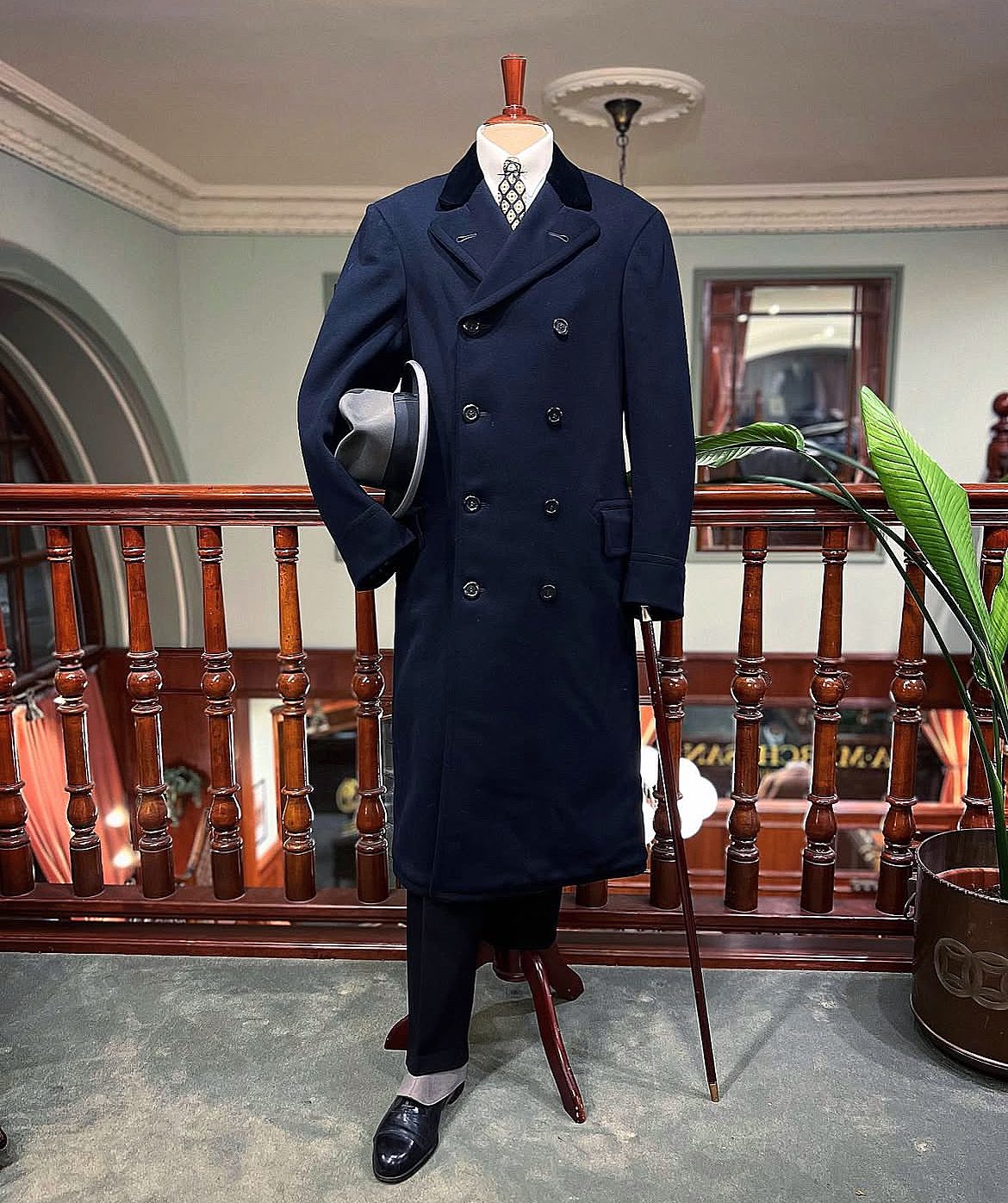 Mannequin wearing a navy double-breasted overcoat with a black velvet collar, white shirt, patterned tie, holding a grey fedora and a walking stick.