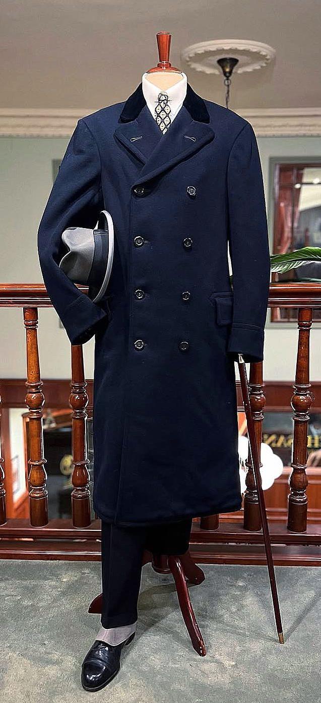 Mannequin wearing a navy double-breasted overcoat with a black velvet collar, white shirt, patterned tie, holding a grey fedora and a walking stick.