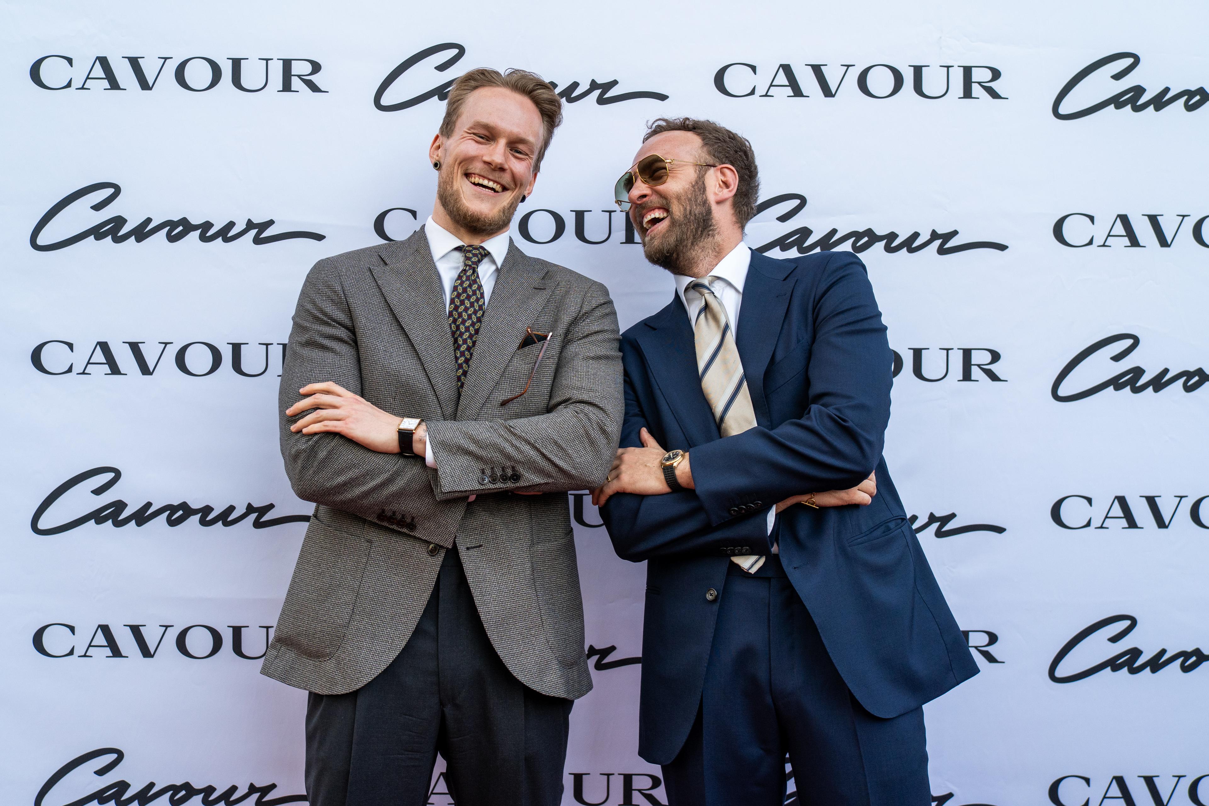 Two men in suits laughing with crossed arms in front of a white backdrop with the Cavour logo.