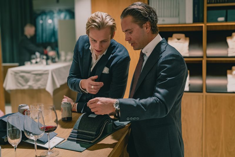 Two men in suits examine fabric swatches on a wooden counter with drinks nearby.
