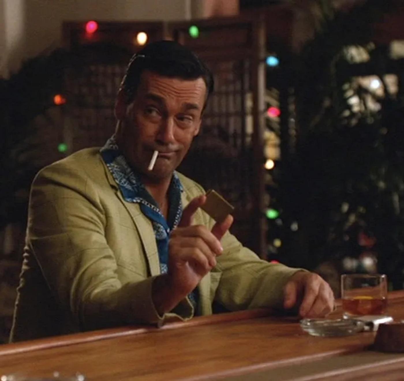 A man in a light green blazer and blue patterned shirt winks, holding a lighter with a cigarette in his mouth, at a bar.